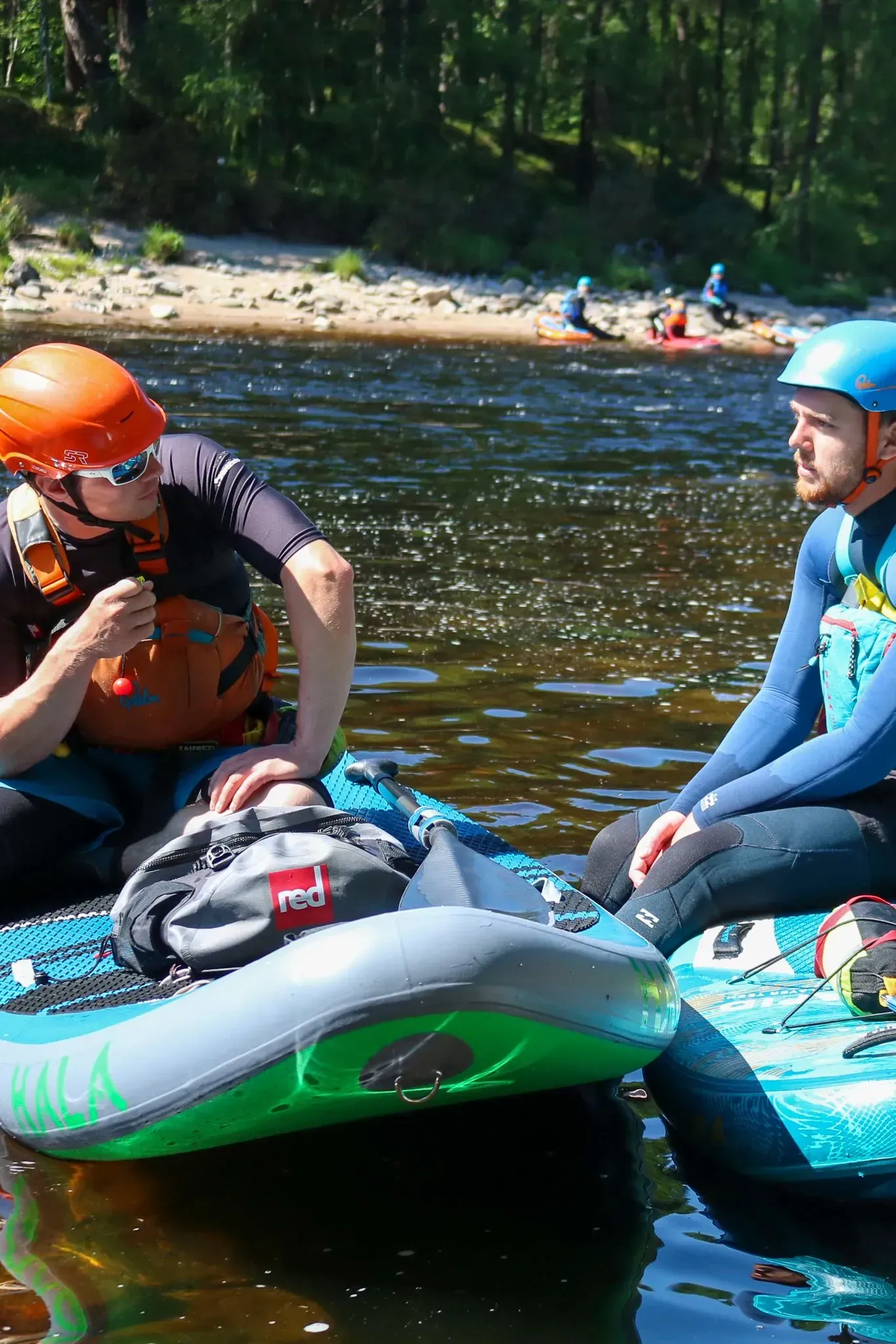 Two people in safety gear sit on paddleboards in a river, talking. One wears an orange helmet, the other a blue one.