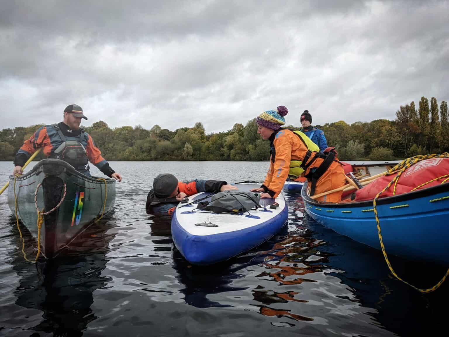 People in life jackets assisting a person in the water. Two canoes and a kayak surround them on a lake with overcast skies.