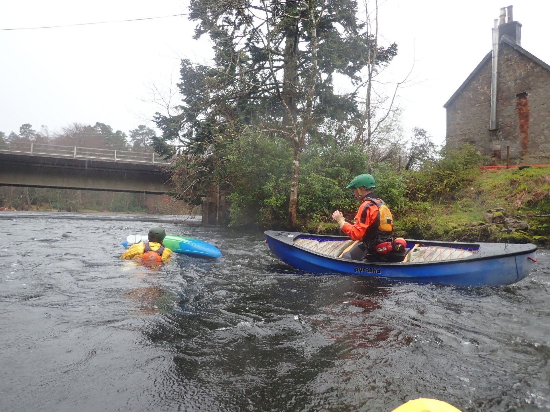 A person in an orange life jacket and a canoe assists another in the water near a bridge and building on a river.