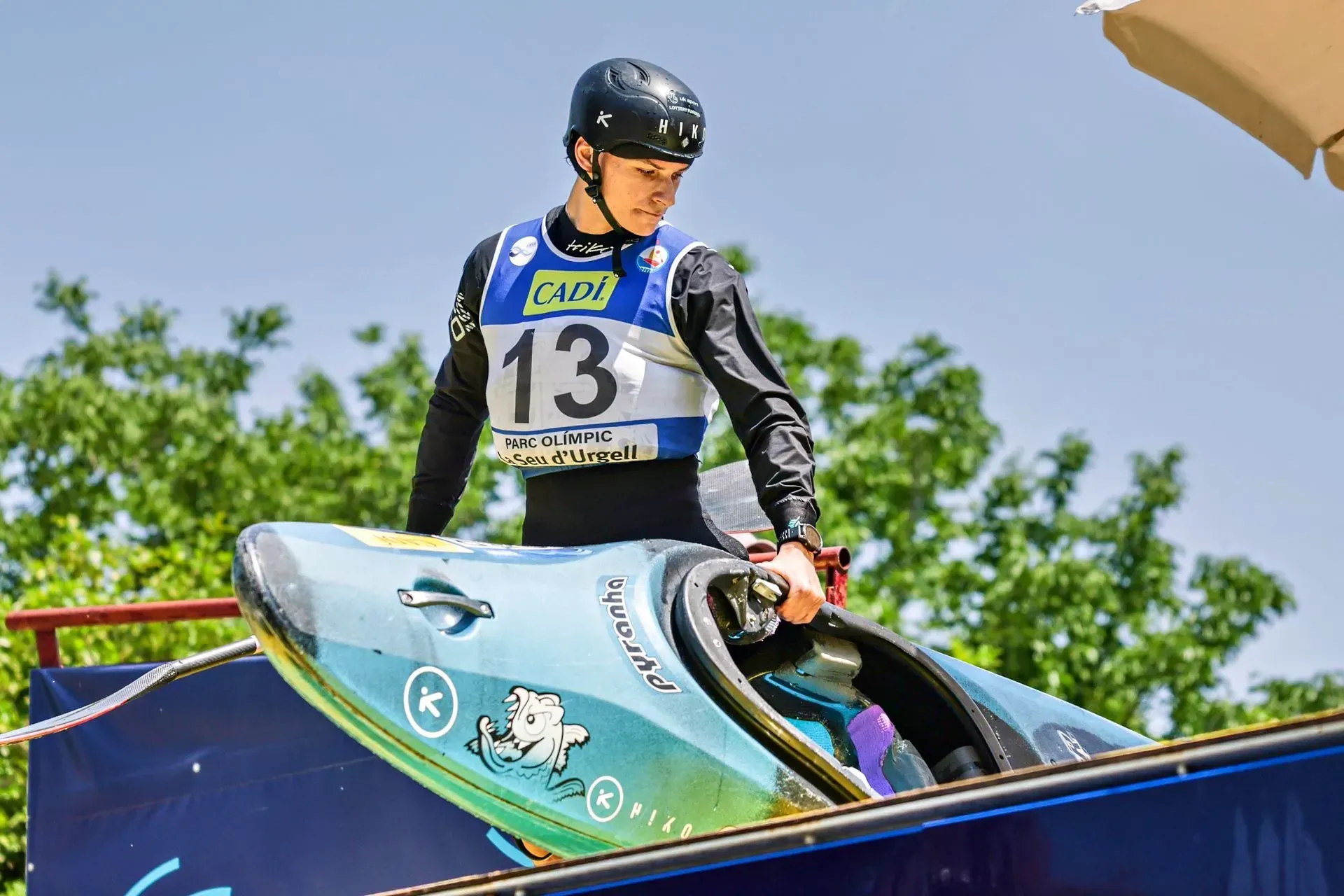 A kayaker in a black helmet and jersey holds a colorful kayak, preparing to enter the water. Number 13 on his jersey.