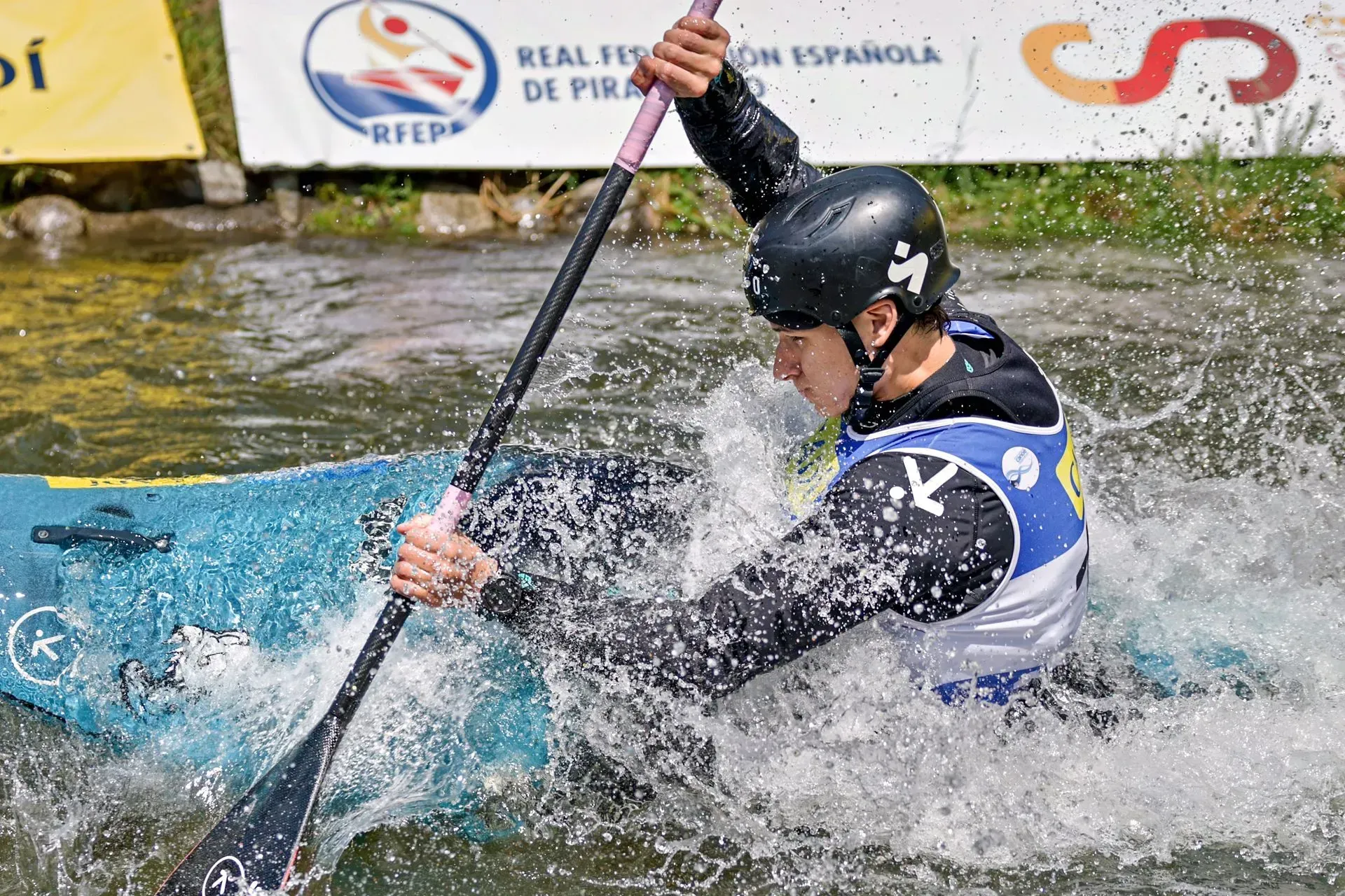 A kayaker in a black and white uniform paddles vigorously through splashing water during a race.