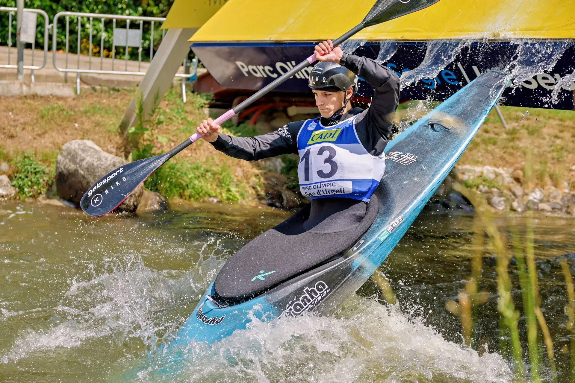 A kayaker in a blue and black boat navigates whitewater, holding a paddle, during a race with the number 13 visible on their bib.