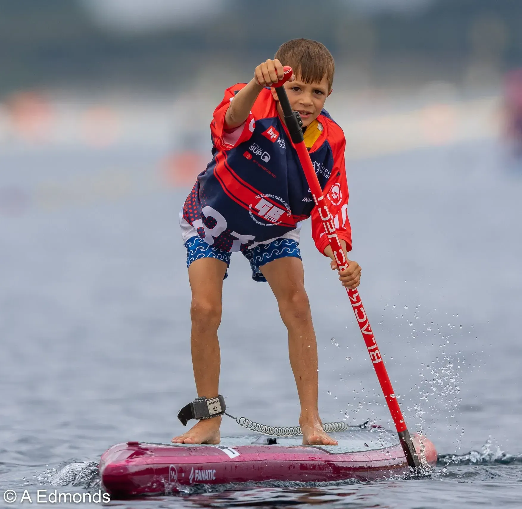 Young boy on a stand-up paddleboard in open water, wearing a red and blue jersey, pointing forward with his paddle.