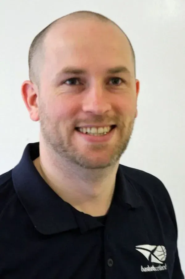 A fair-skinned man with a shaved head smiles while wearing a dark blue polo shirt, standing in front of a white background.