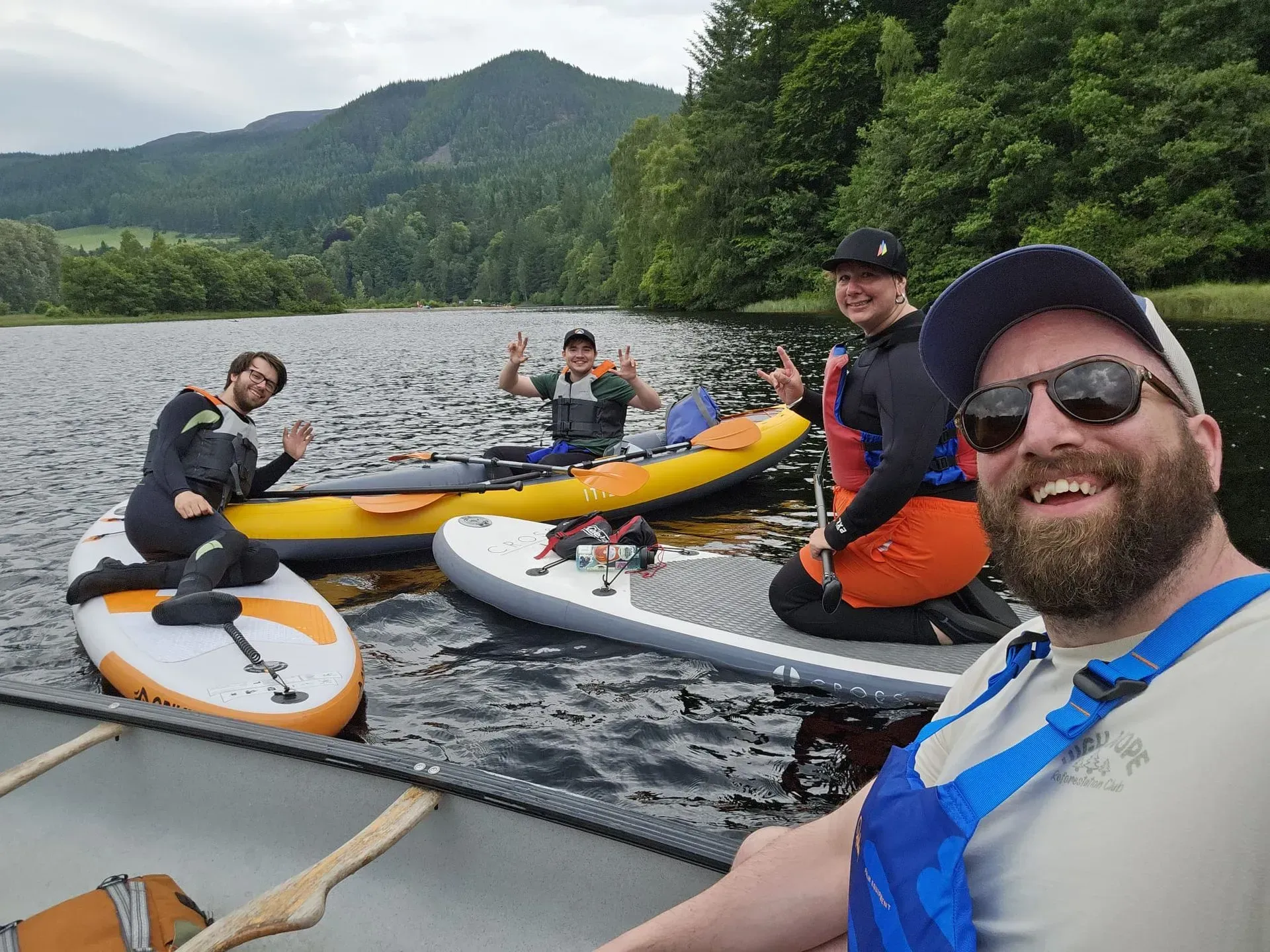Group of friends paddleboarding and kayaking on a lake, smiling and giving peace signs. Mountains and trees in the background.