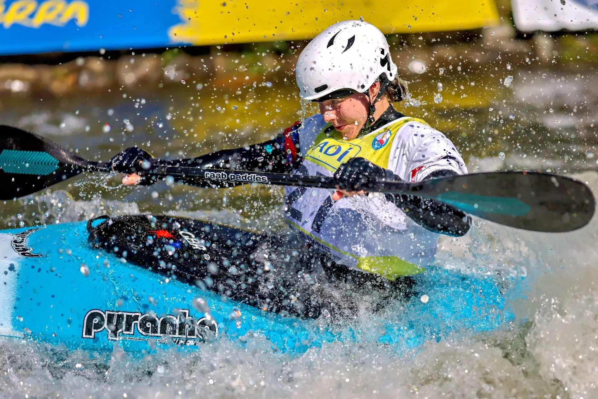 A kayaker in a turquoise kayak navigating whitewater rapids, paddles raised, wearing a white helmet.