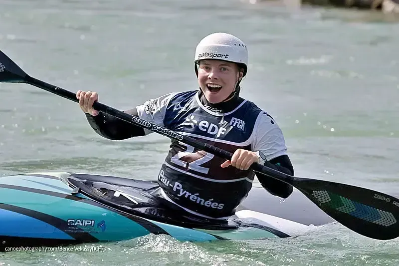 Kayaker in white helmet, smiling, paddling in a turquoise kayak on whitewater. The person wears a navy and white competition jersey, with a backdrop of water and sky.