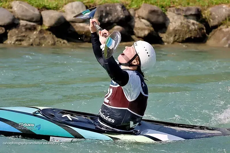 A kayaker raises a paddle in triumph after navigating a whitewater course. They wear a helmet and wetsuit, in a turquoise and white kayak.