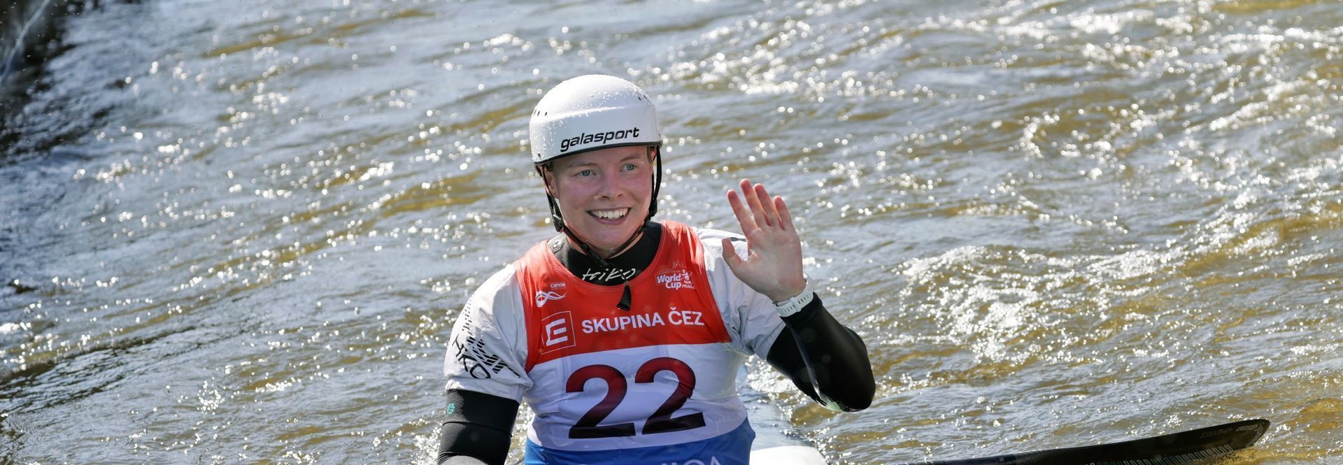 A female kayaker wearing a white helmet waves, smiling. She is in a river during a competition, wearing a red and blue jersey with the number 22.