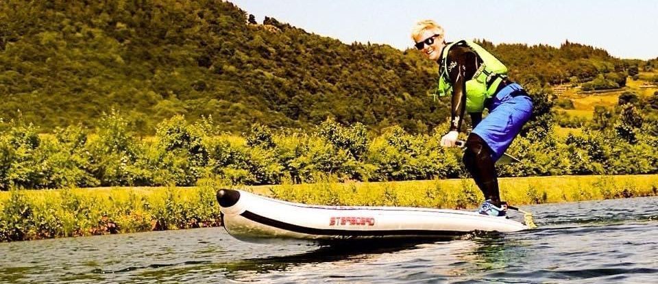 A person in sunglasses and a life vest stands on a motorized inflatable watercraft, navigating a river with a forested background.