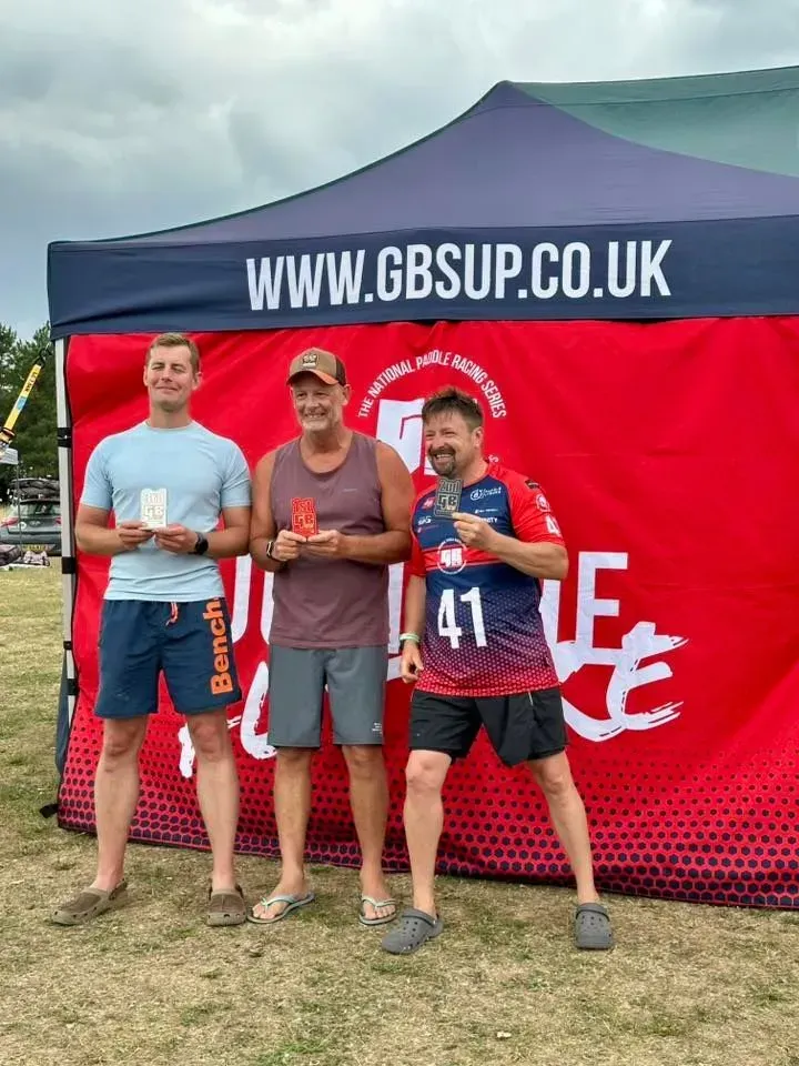 Three men stand in front of a red banner, holding trophies. They appear to be winners at an outdoor event.