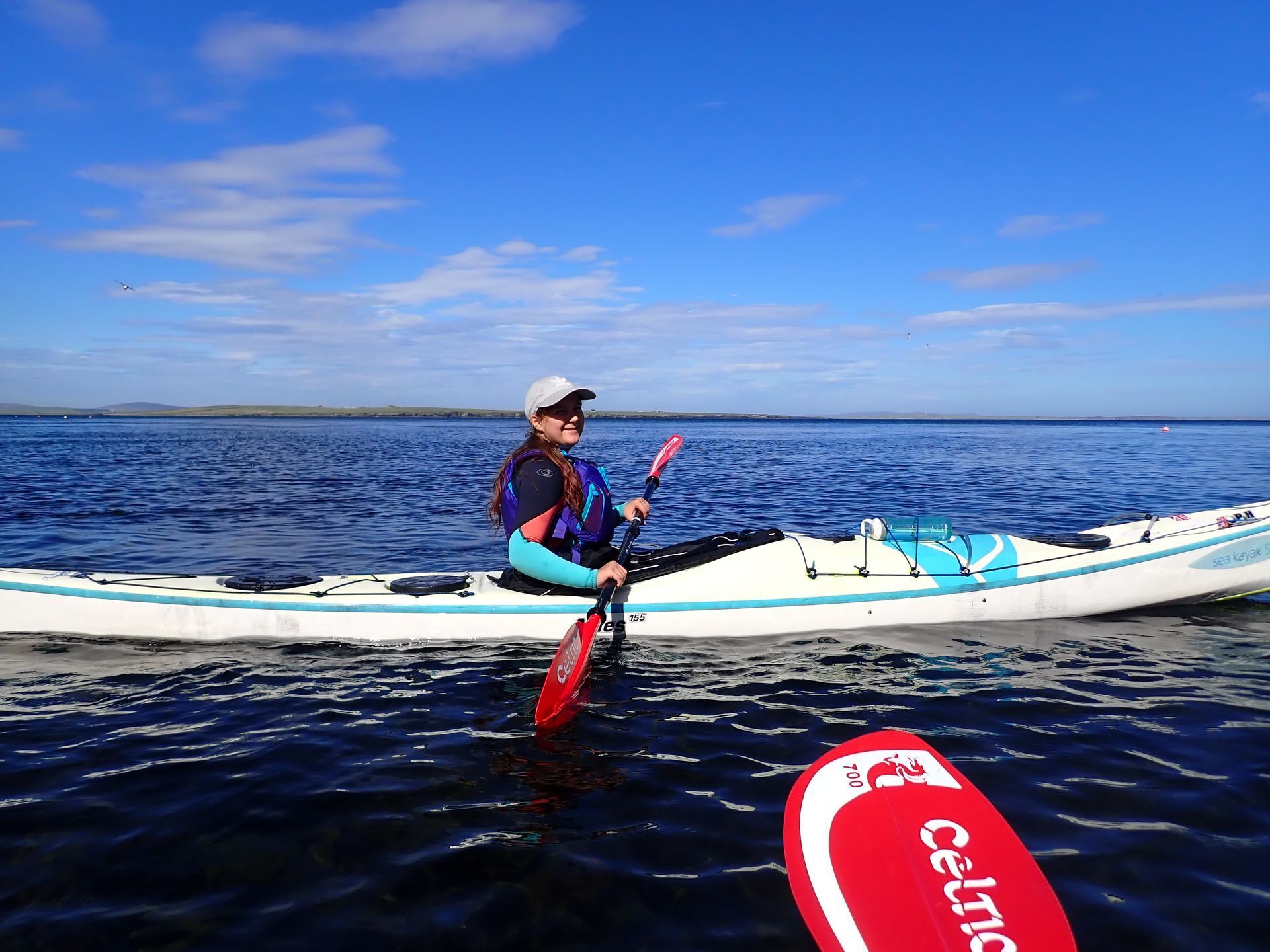 Person kayaking on a calm blue lake under a bright blue sky, holding a paddle, with a second paddle in the foreground.
