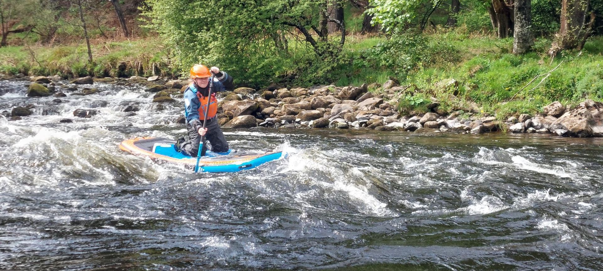 A person on a stand-up paddleboard navigates a rushing river. They wear a helmet, orange life vest, and are paddling.