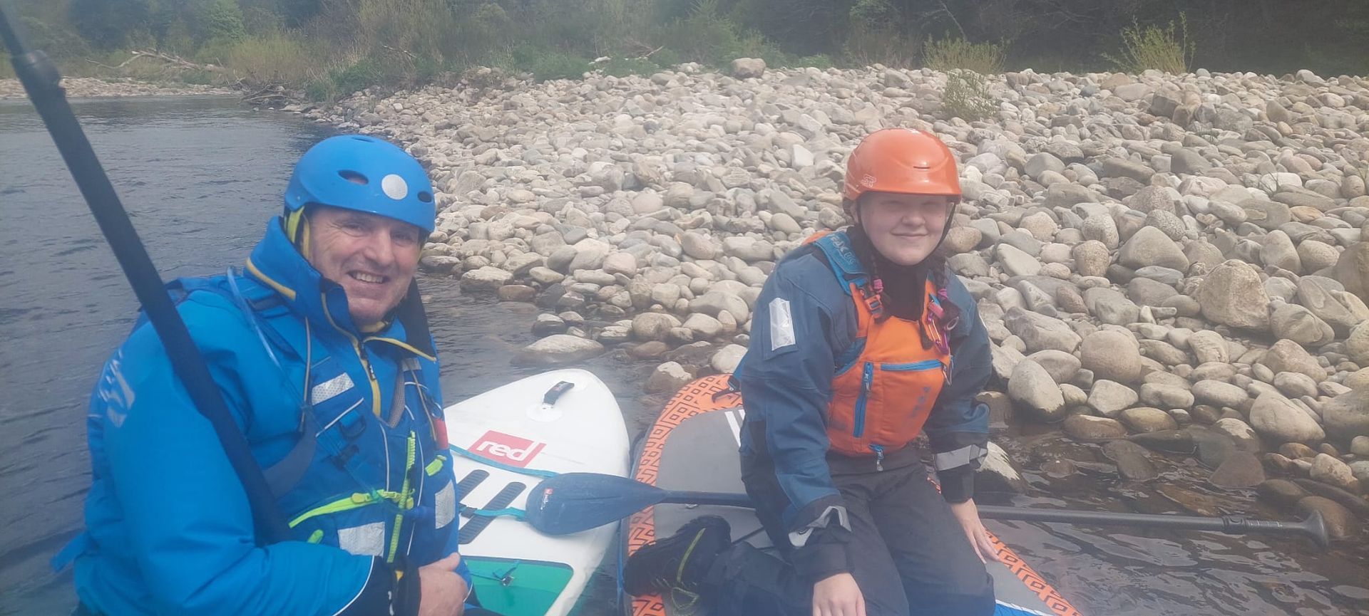 Two people in helmets and life vests smile while rafting on a river with a rocky bank.
