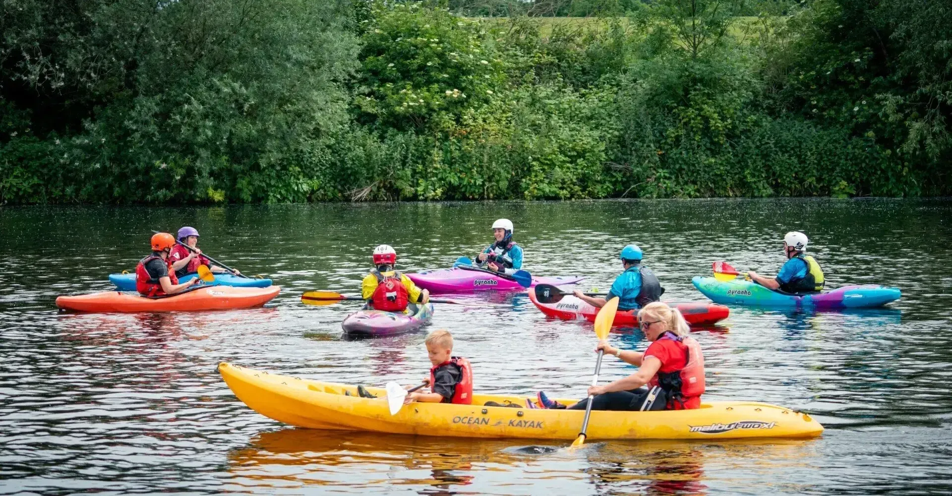 People kayaking on a calm river. Several kayaks of different colors with paddlers wearing life vests are on the water, surrounded by greenery.