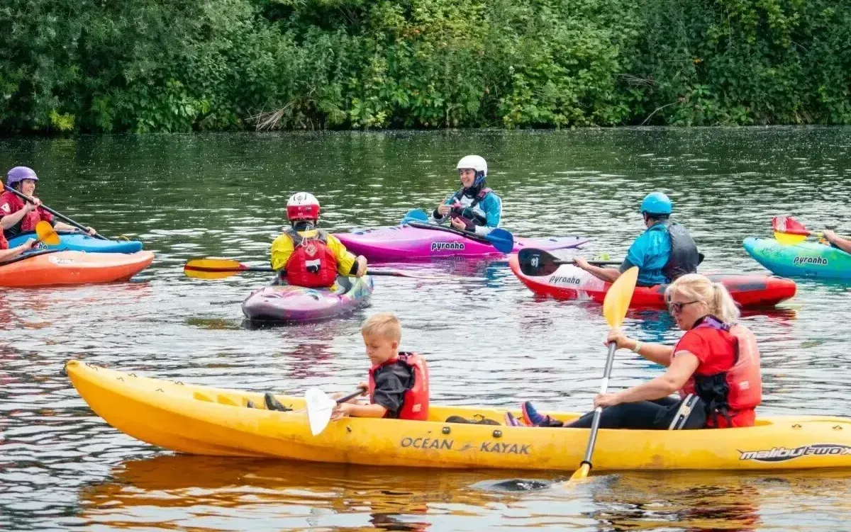 Group of people kayaking on a calm river. Several colorful kayaks, various ages wearing life vests and helmets, enjoying the water in a natural setting.