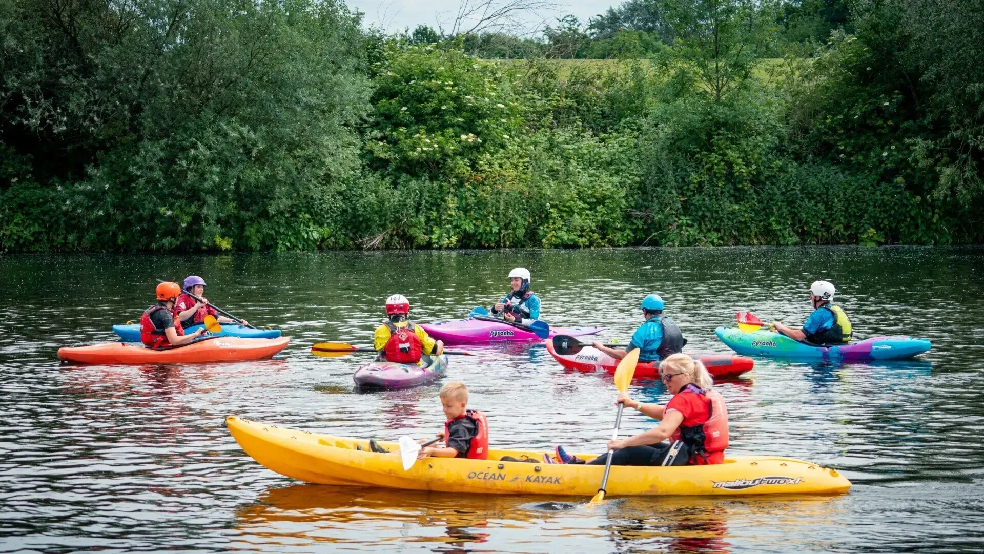 A group of people in kayaks on a river. They wear life vests and helmets, paddling near a green, tree-lined bank.