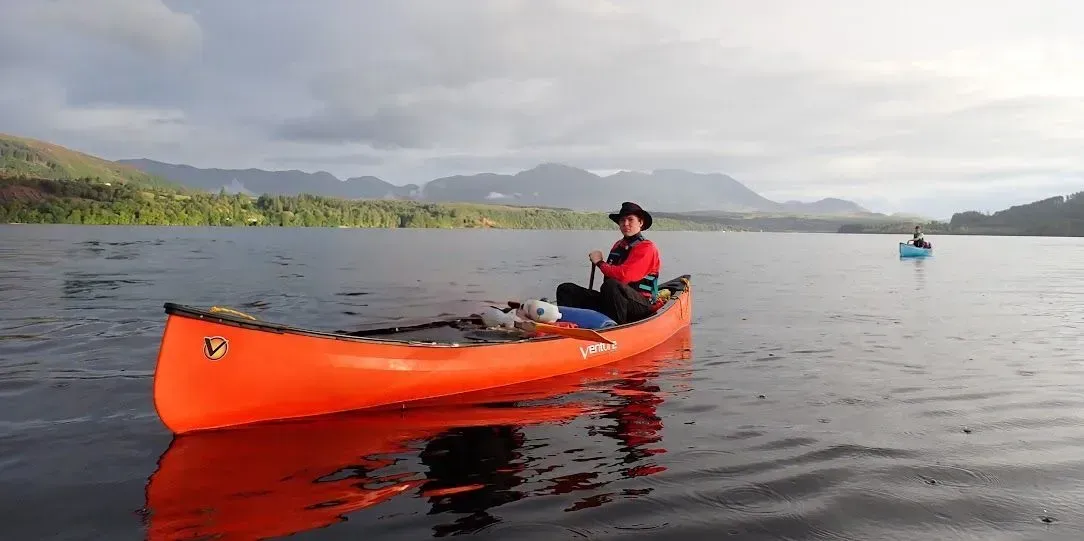 A person canoes on calm water, with mountains in the background. The canoe is bright orange, reflecting on the water.