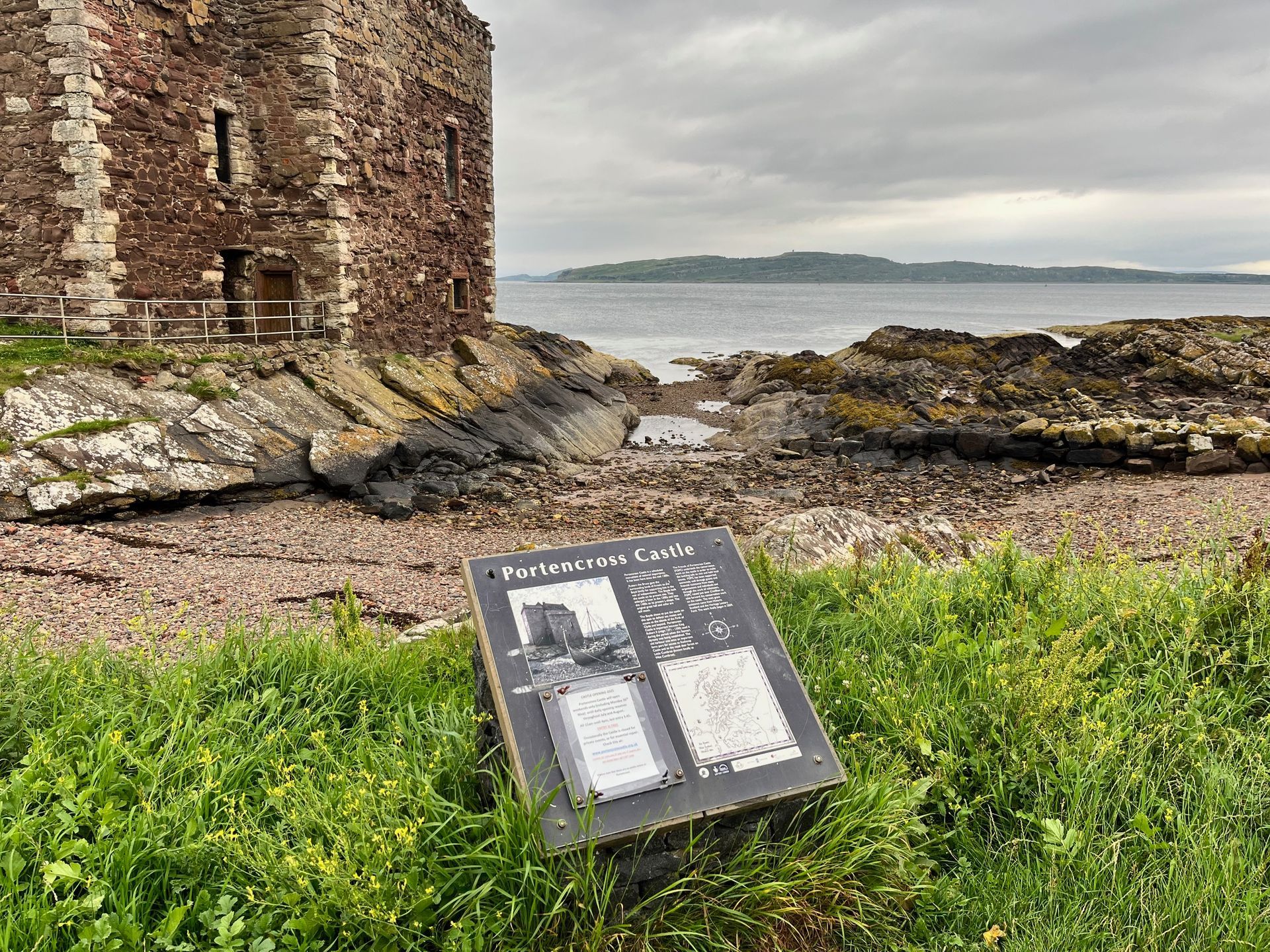 Ruined castle beside a rocky shore with an informational sign in front. Cloudy sky and distant island.