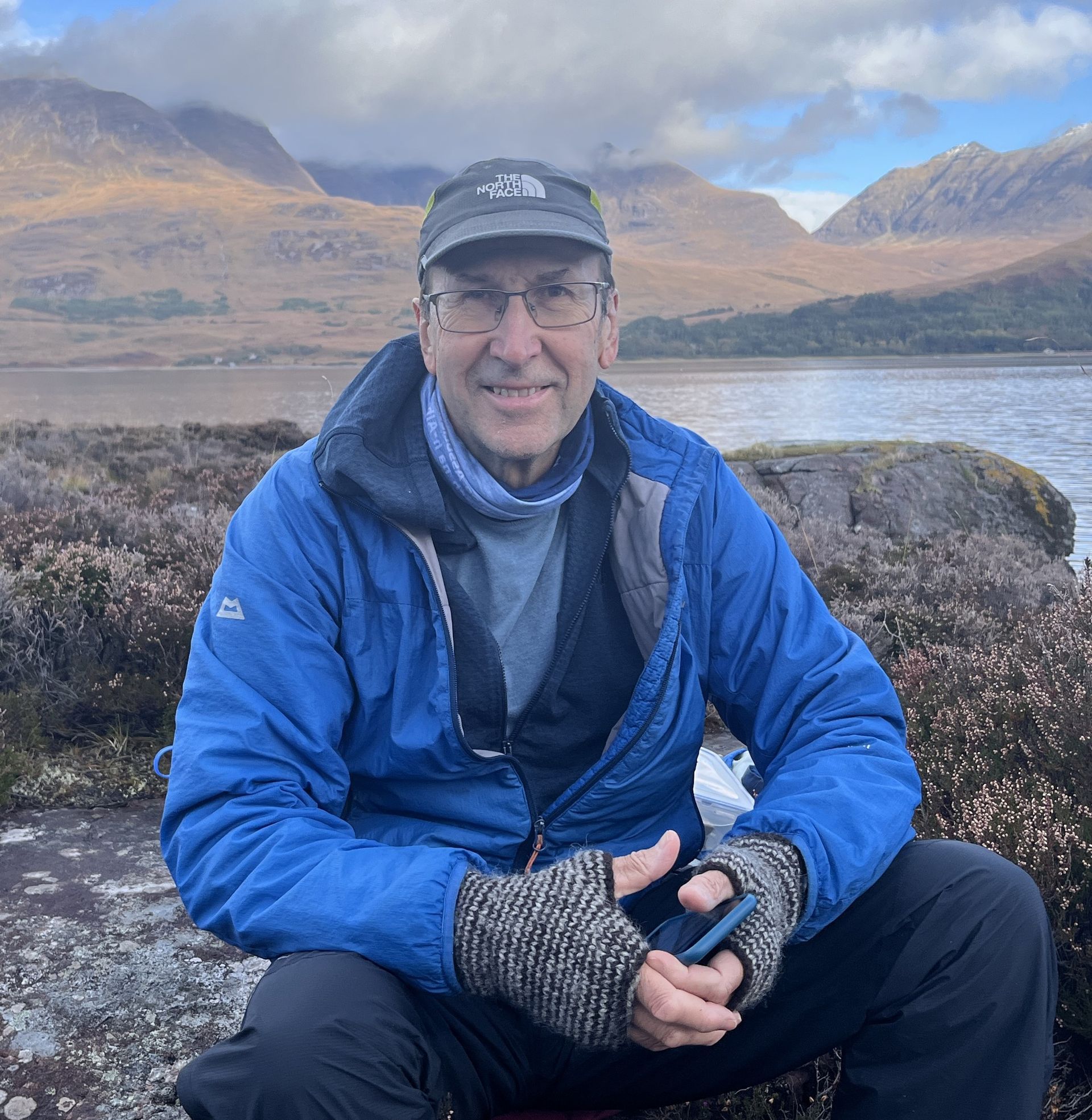 Man in blue jacket and cap, smiling at the camera, in front of a lake and mountains. He's wearing fingerless gloves and holding a phone.