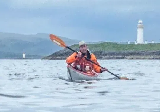 Man kayaking on water near a small island with a white lighthouse. He's wearing an orange jacket and using a paddle.