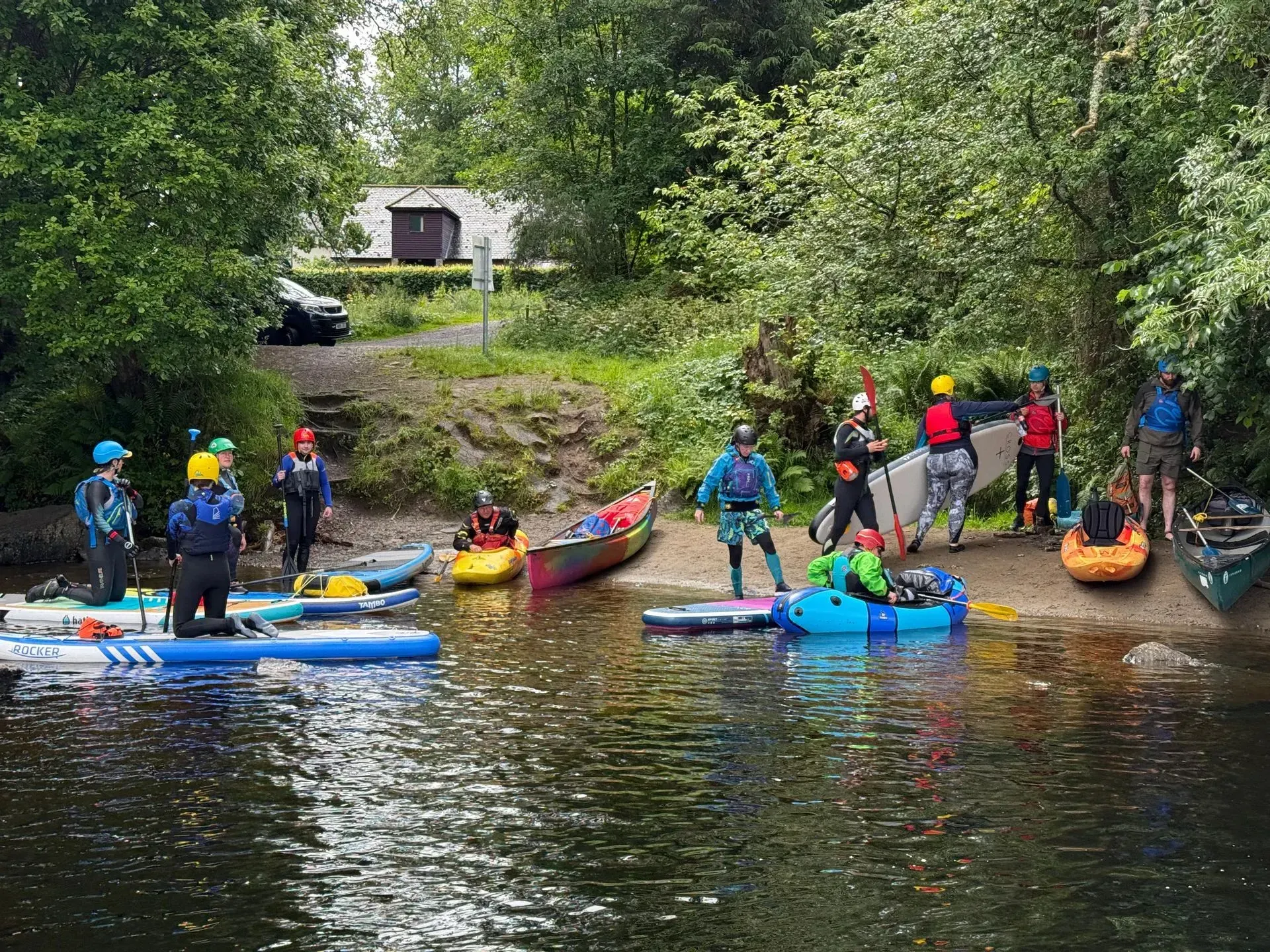 People in water gear prepare paddleboards, kayaks, and a canoe at a riverbank, with trees and a small building in the background.