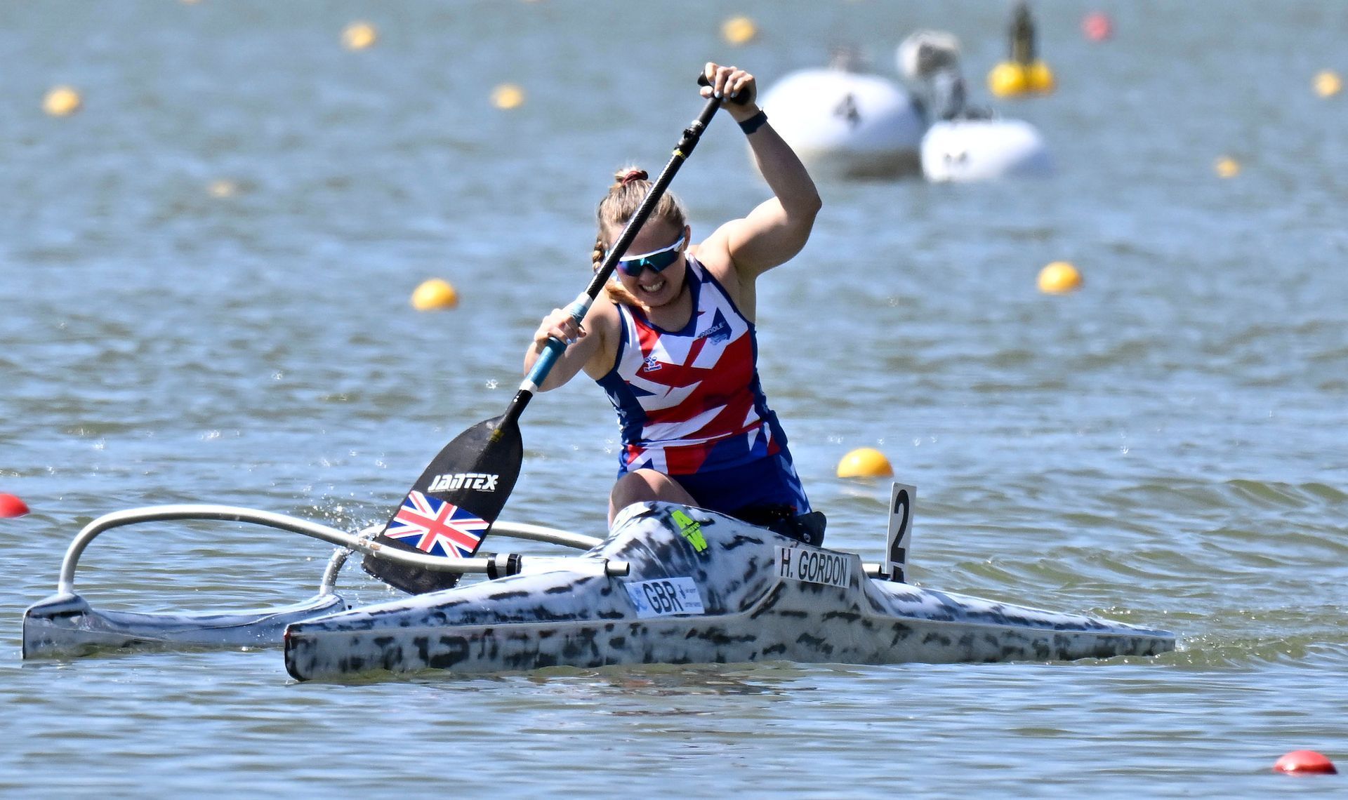 A female para-canoeist paddles a gray canoe, wearing a British flag top, with her right arm raised in celebration. They are in a body of water.
