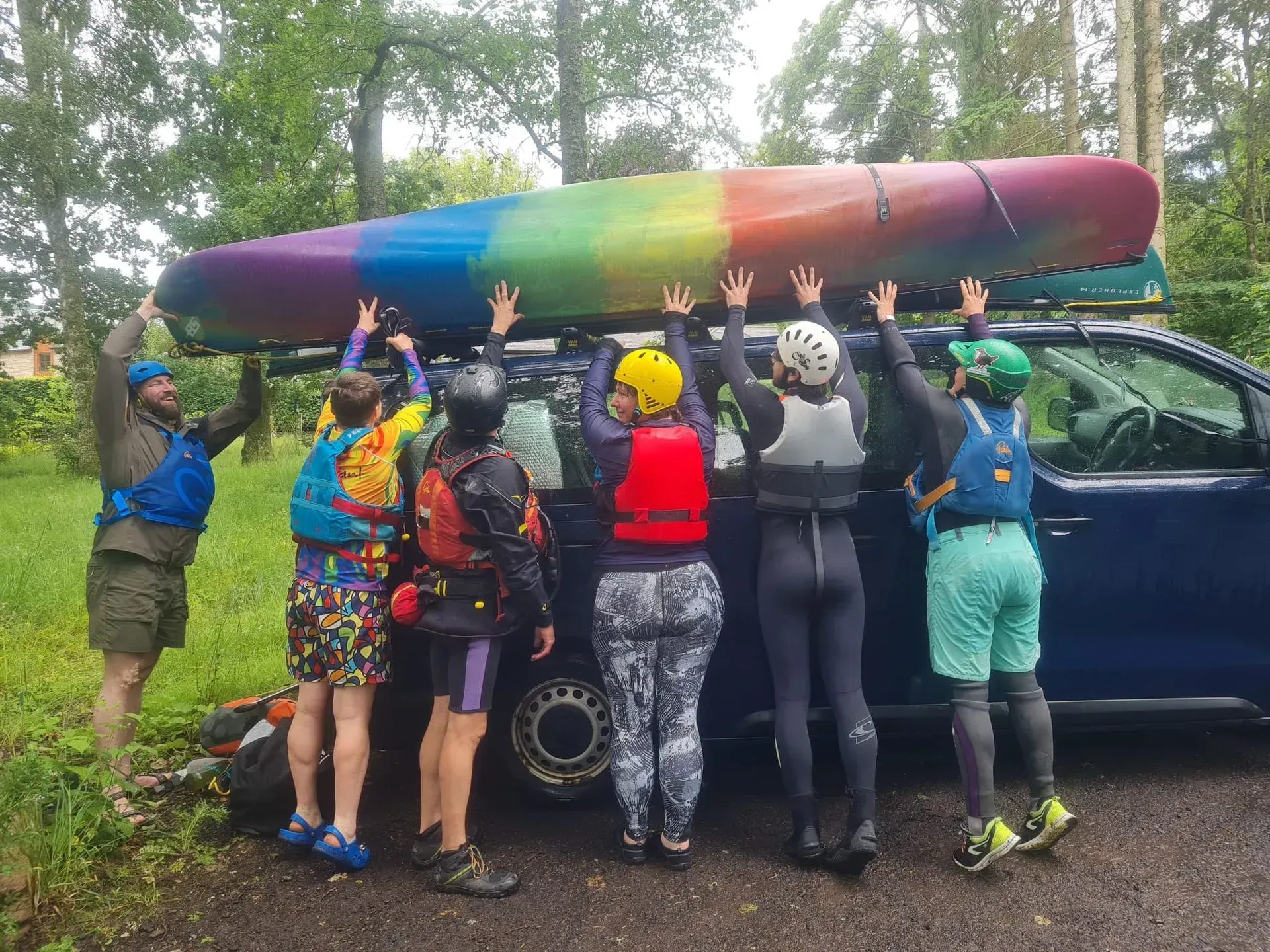 A group of people lift a rainbow-colored canoe onto the roof of a blue car. They're outdoors, wearing life vests and gear.