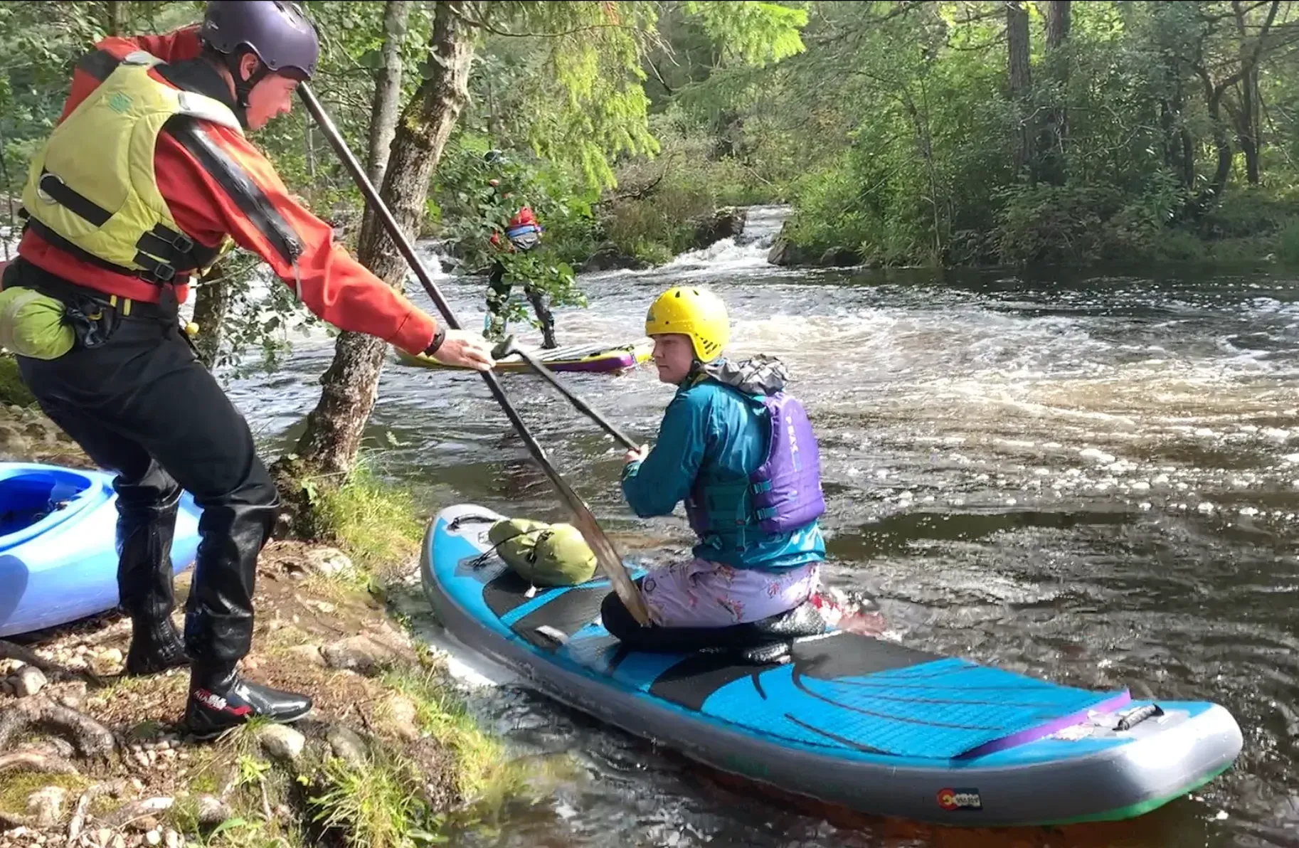 A person on a blue paddleboard is assisted by another person in a river, near a wooded bank. Both wear helmets and life vests.
