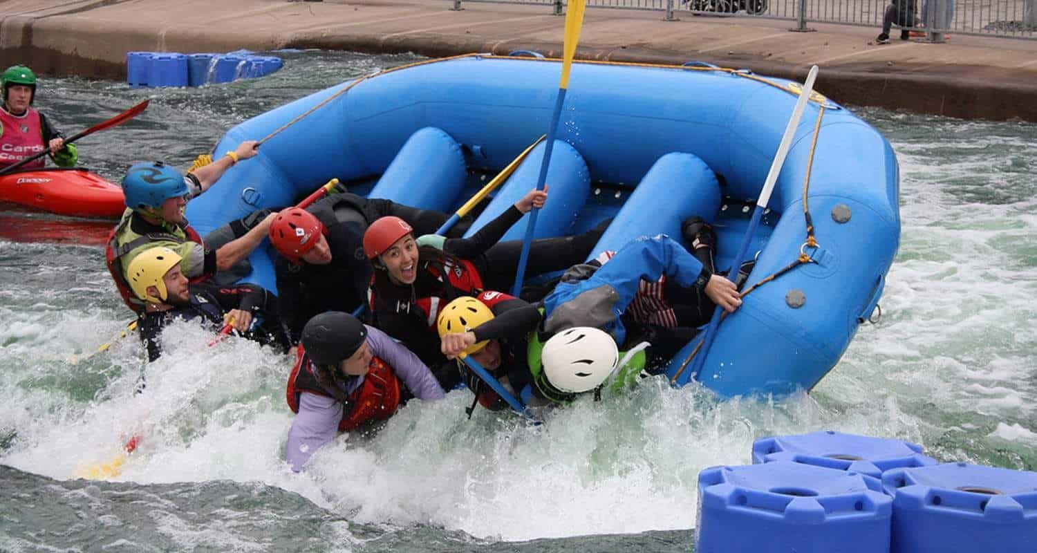 Rafting crew capsizes in whitewater rapids. Several people wearing helmets and life vests are in the water and struggling with the overturned raft.