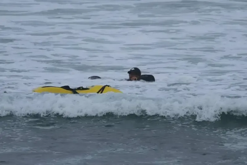 A person in a black wetsuit in the ocean with a yellow rescue buoy. The person is near breaking waves on an overcast day.