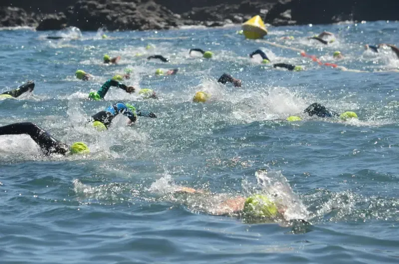 Swimmers in wetsuits in open water race, churning up the blue waves. Yellow buoys mark the course near a rocky coastline.