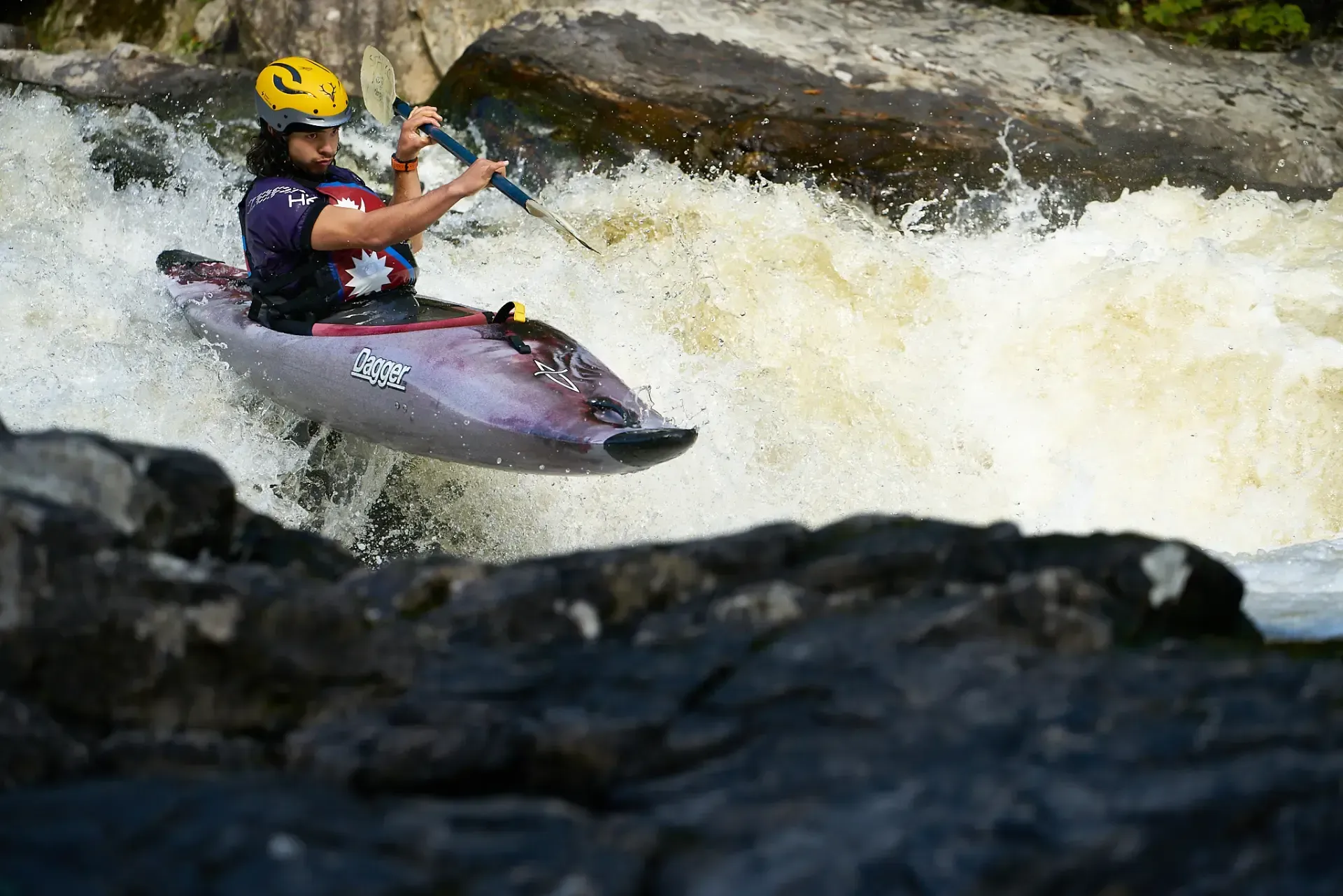 A kayaker in a purple boat navigates a waterfall, paddling through white water. They wear a yellow helmet and a purple vest.