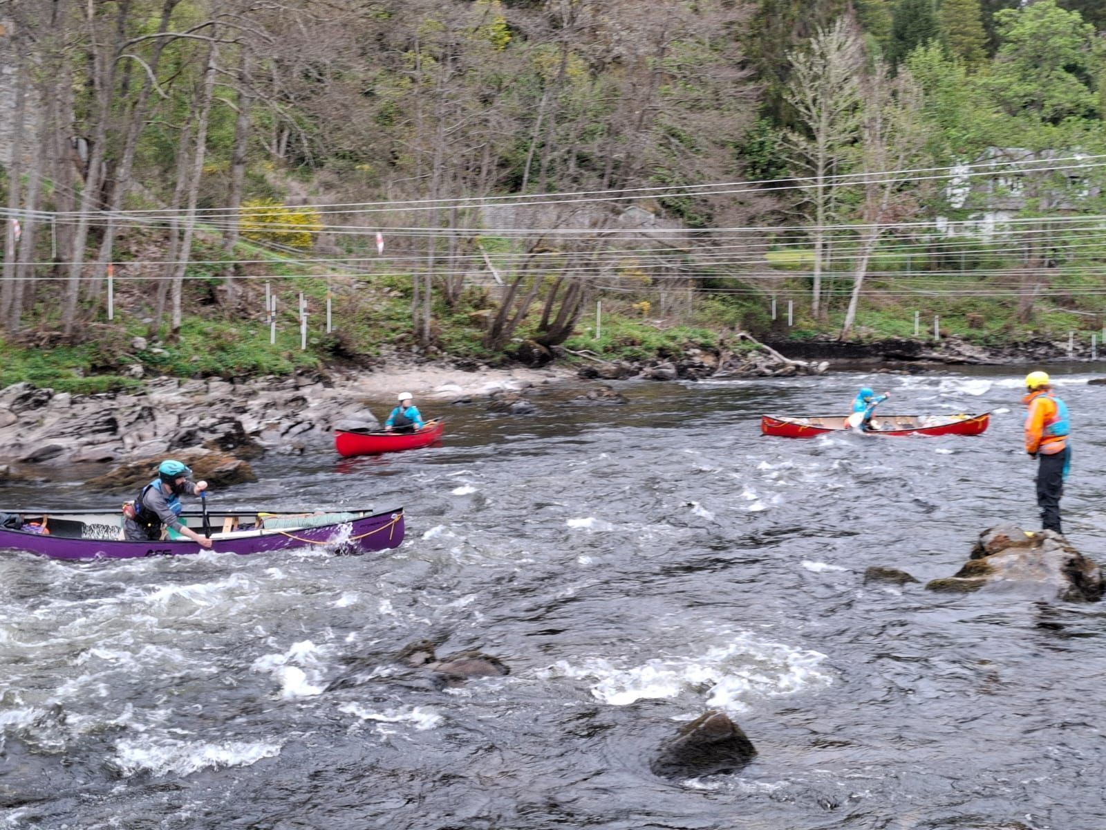 Canoes navigate a fast-flowing river, with people in the boats wearing helmets and life vests. A person watches from the riverbank.