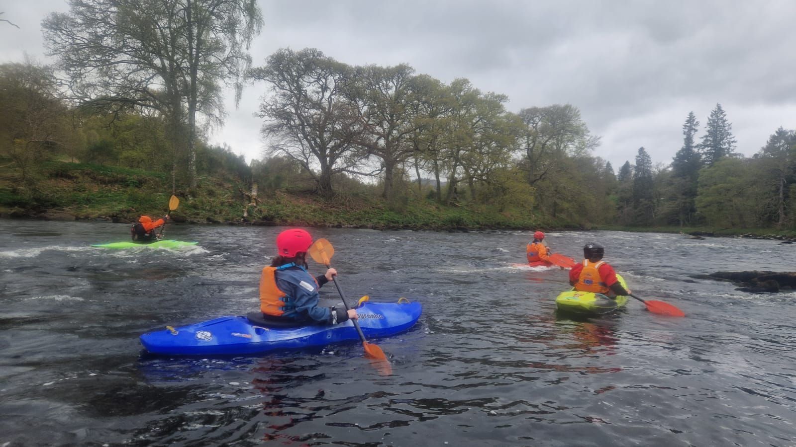 Kayakers in colorful boats paddle down a river on a cloudy day. Trees line the banks.