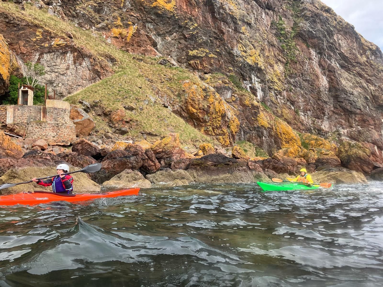 Kayakers near a rocky shoreline. One in an orange kayak, the other in green, paddling near a cliff face with moss and a small building.