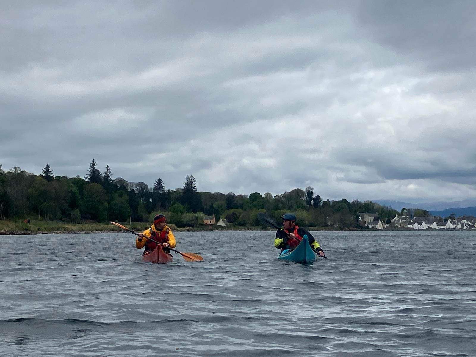 Two kayakers on a lake under an overcast sky. They wear brightly colored life vests and paddle toward the shoreline with trees and buildings.