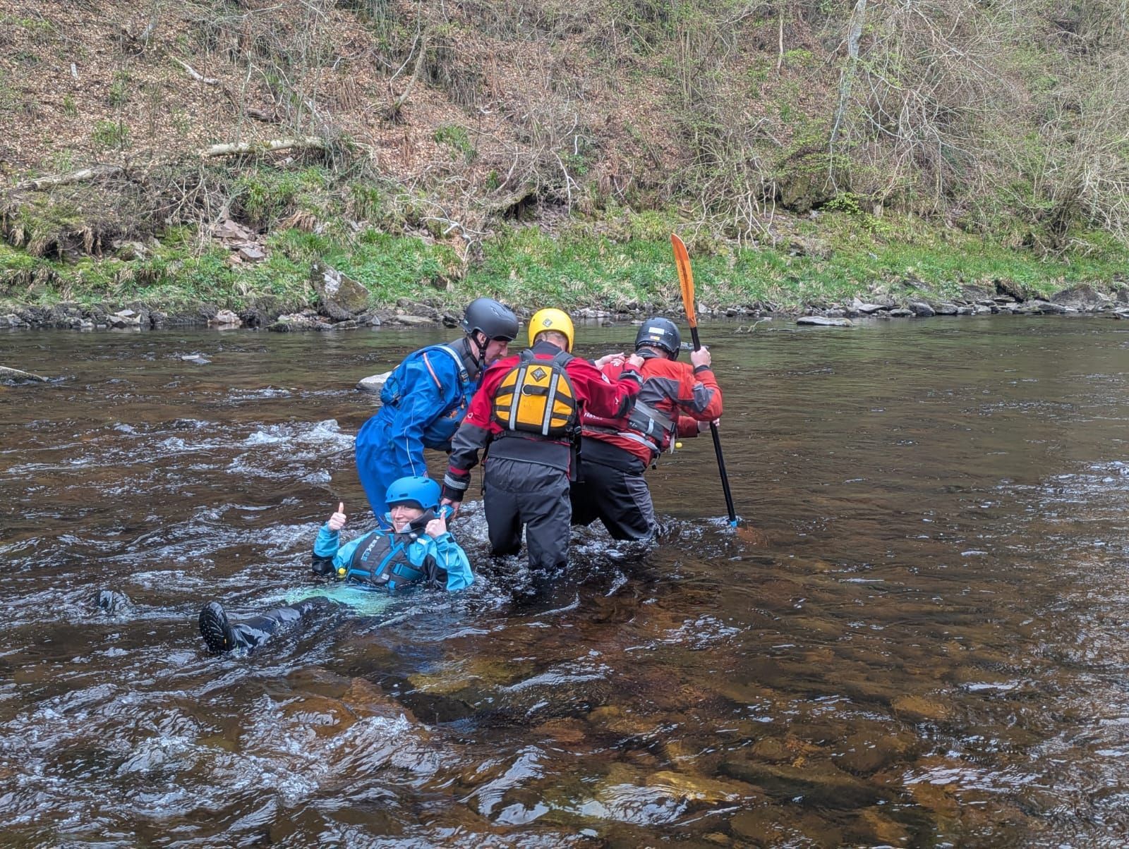 People in the water, assisting someone lying down. They're wearing wetsuits and life vests in a river.