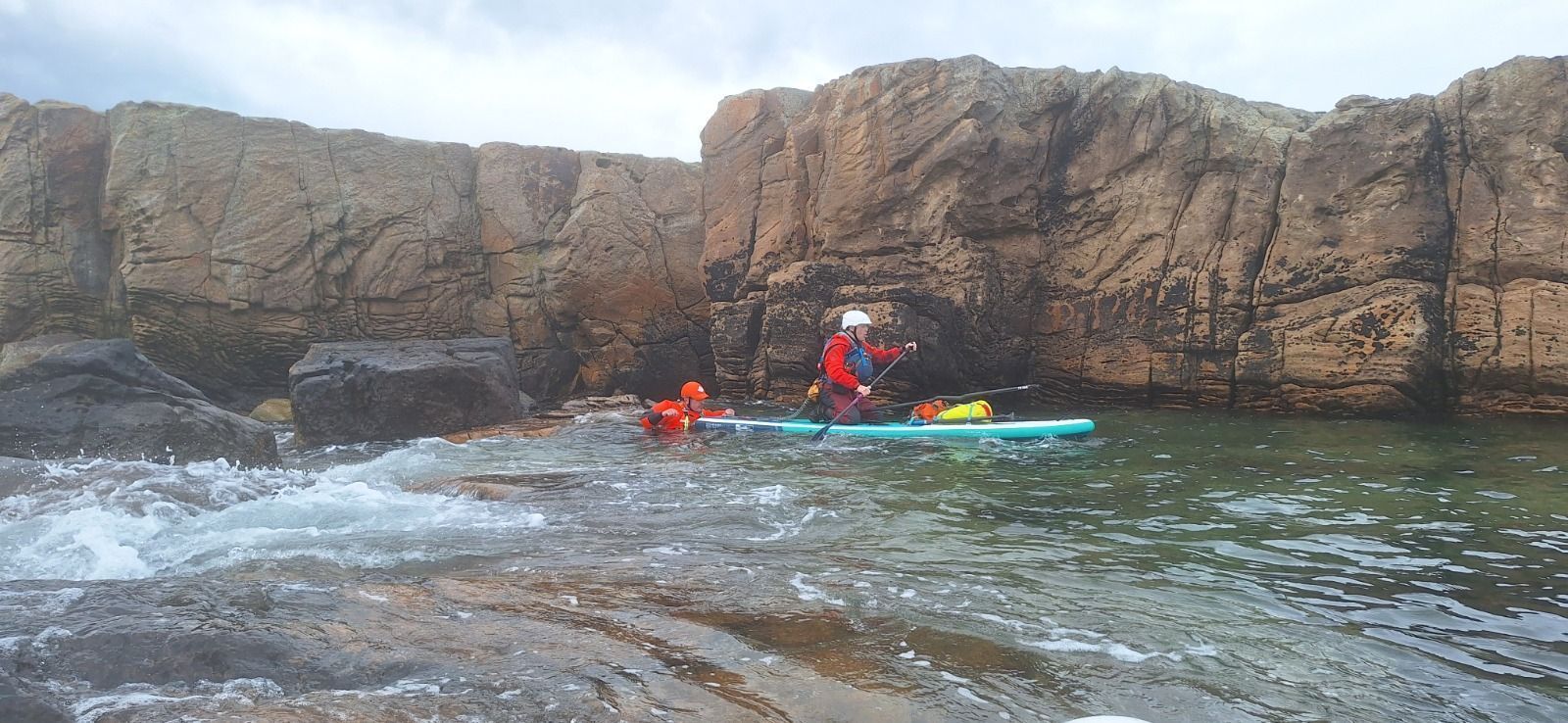 A person in a kayak paddles near large rock formations. Water is choppy, and the sky is overcast.