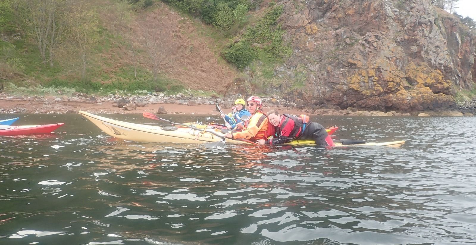 Kayakers on the water near a rocky shore. One person is in the water, being assisted by others in the kayaks.