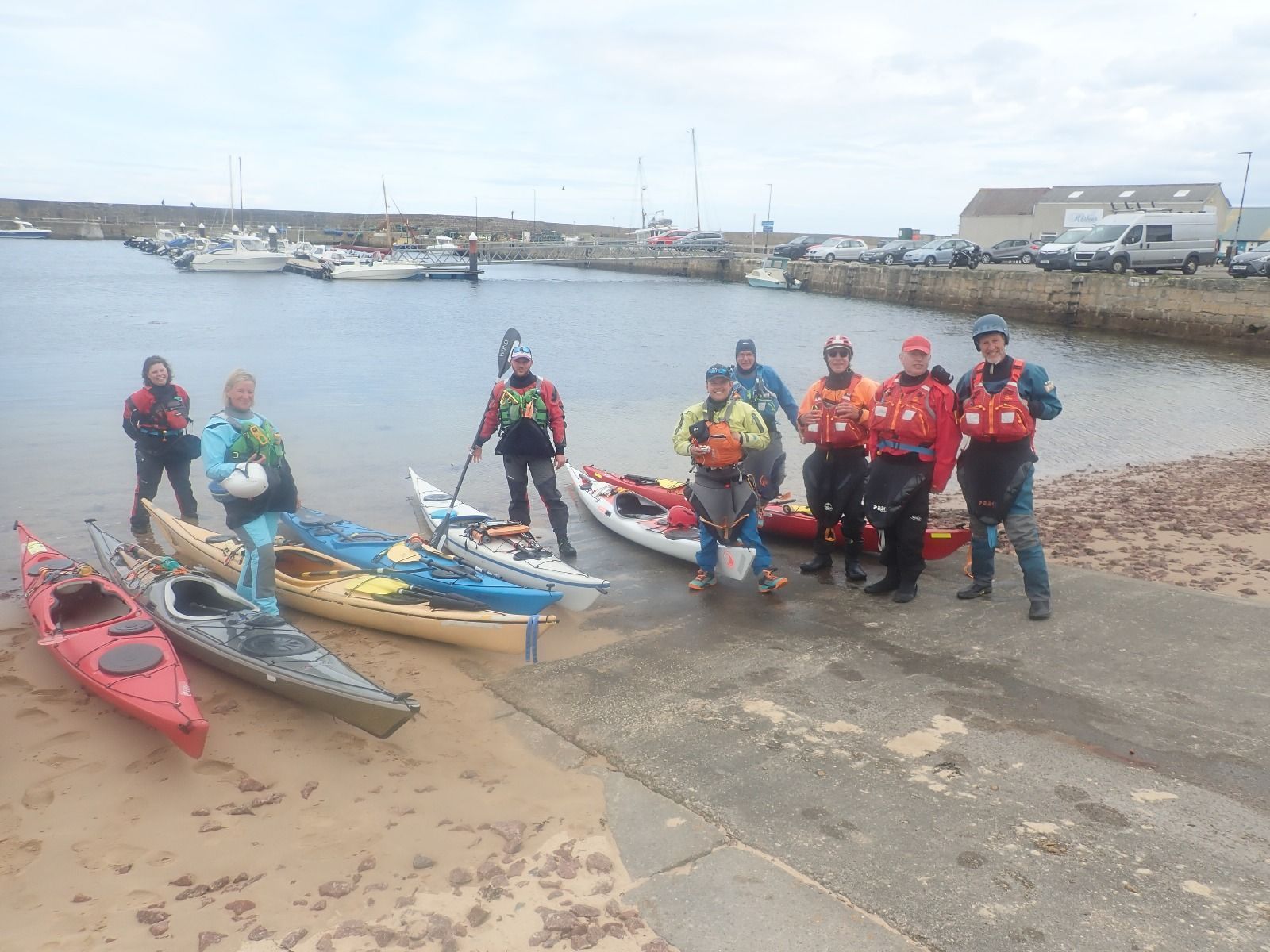 A group of kayakers in bright gear stand with their kayaks on a beach, ready to launch from a harbor.