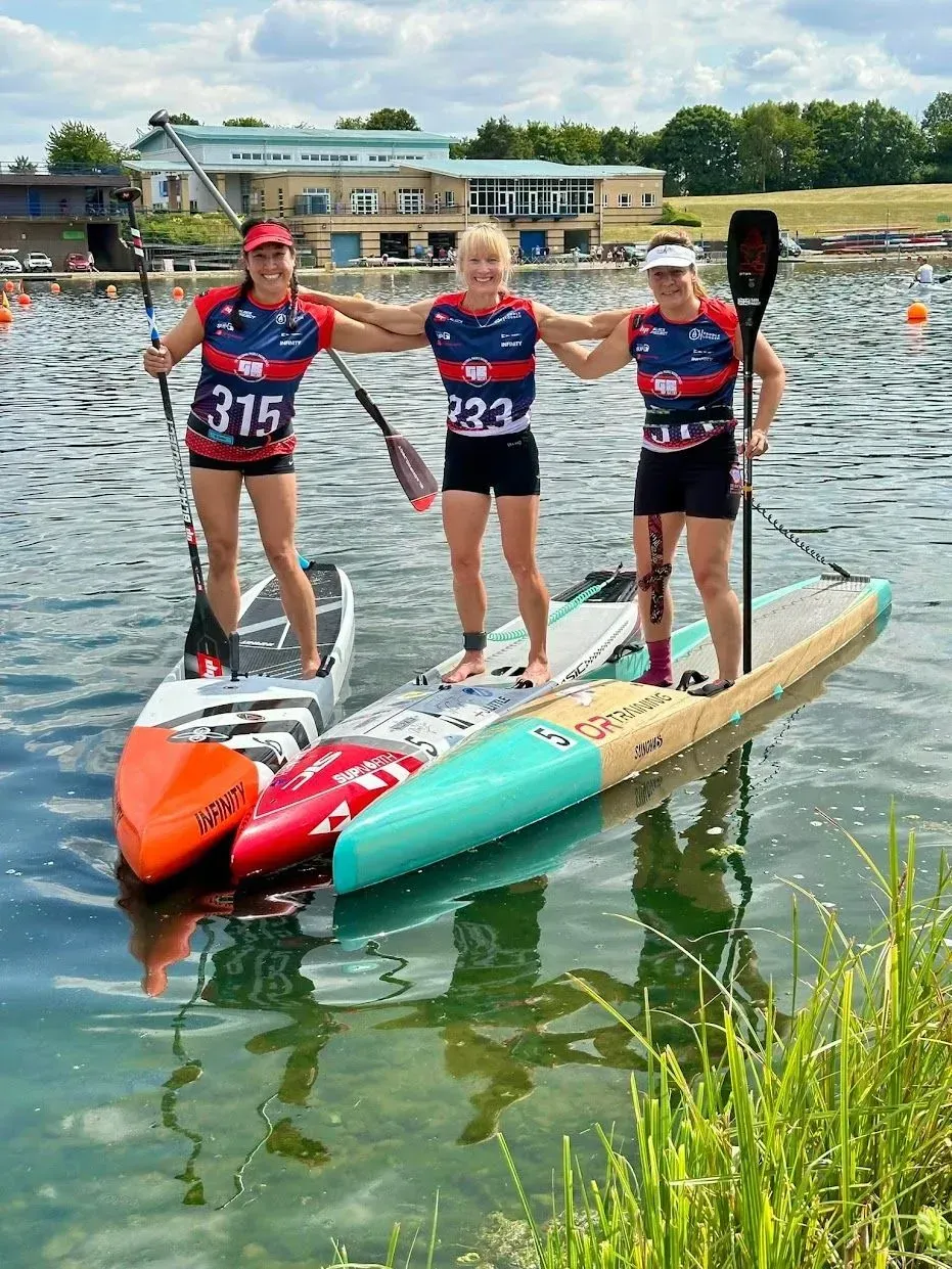 Three women on stand-up paddleboards in a lake, celebrating a race finish. They're wearing matching tops, smiling, and holding paddles.