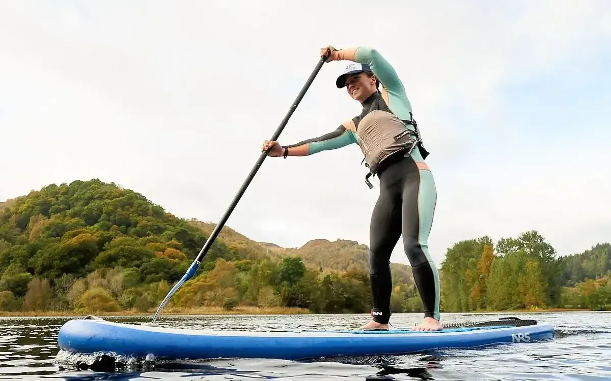 Person on a paddleboard, paddling with a long oar in a lake. They are wearing a wetsuit and life vest.