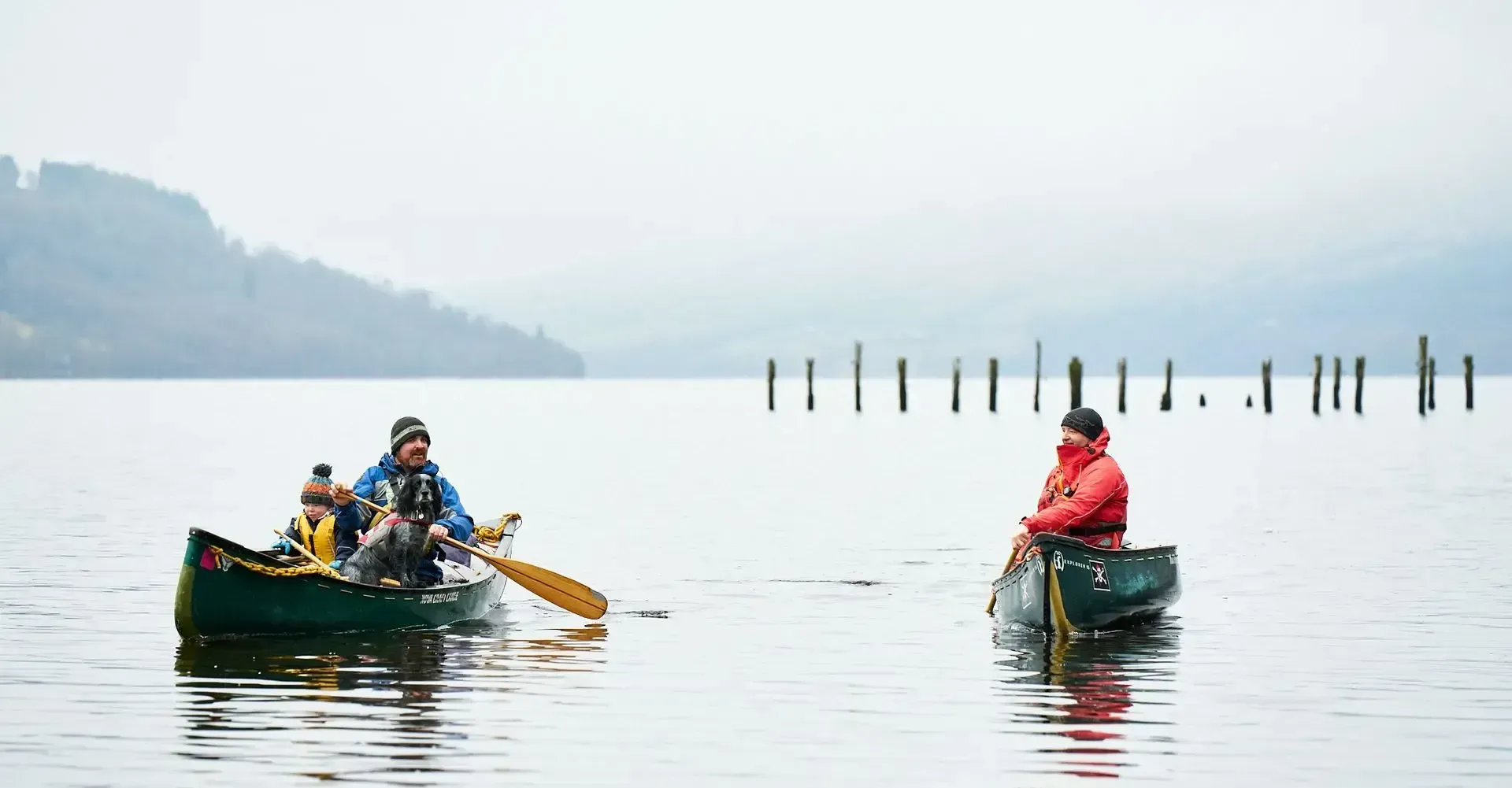 Two canoes on calm water with three people. The person in the canoe on the left paddles. The other canoeist faces them.