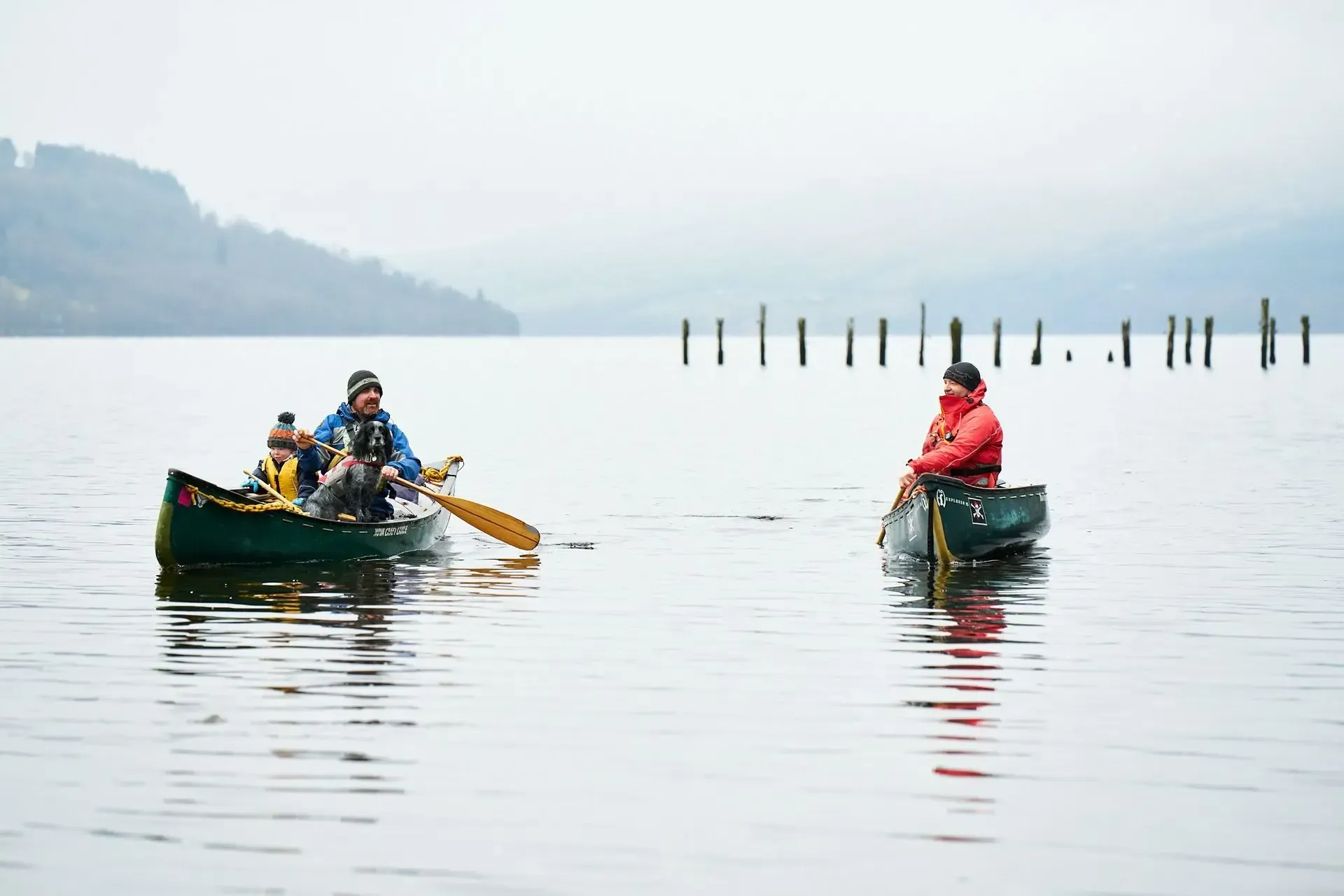 Two canoes on calm water; people in each canoe, paddling. Wooden posts in the distance; overcast day.
