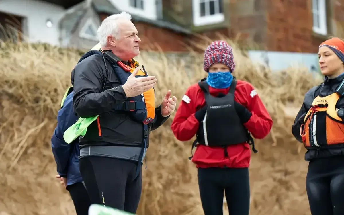 A group of people in cold-weather gear on a sandy beach listen to an older man gesturing, likely before a water activity.