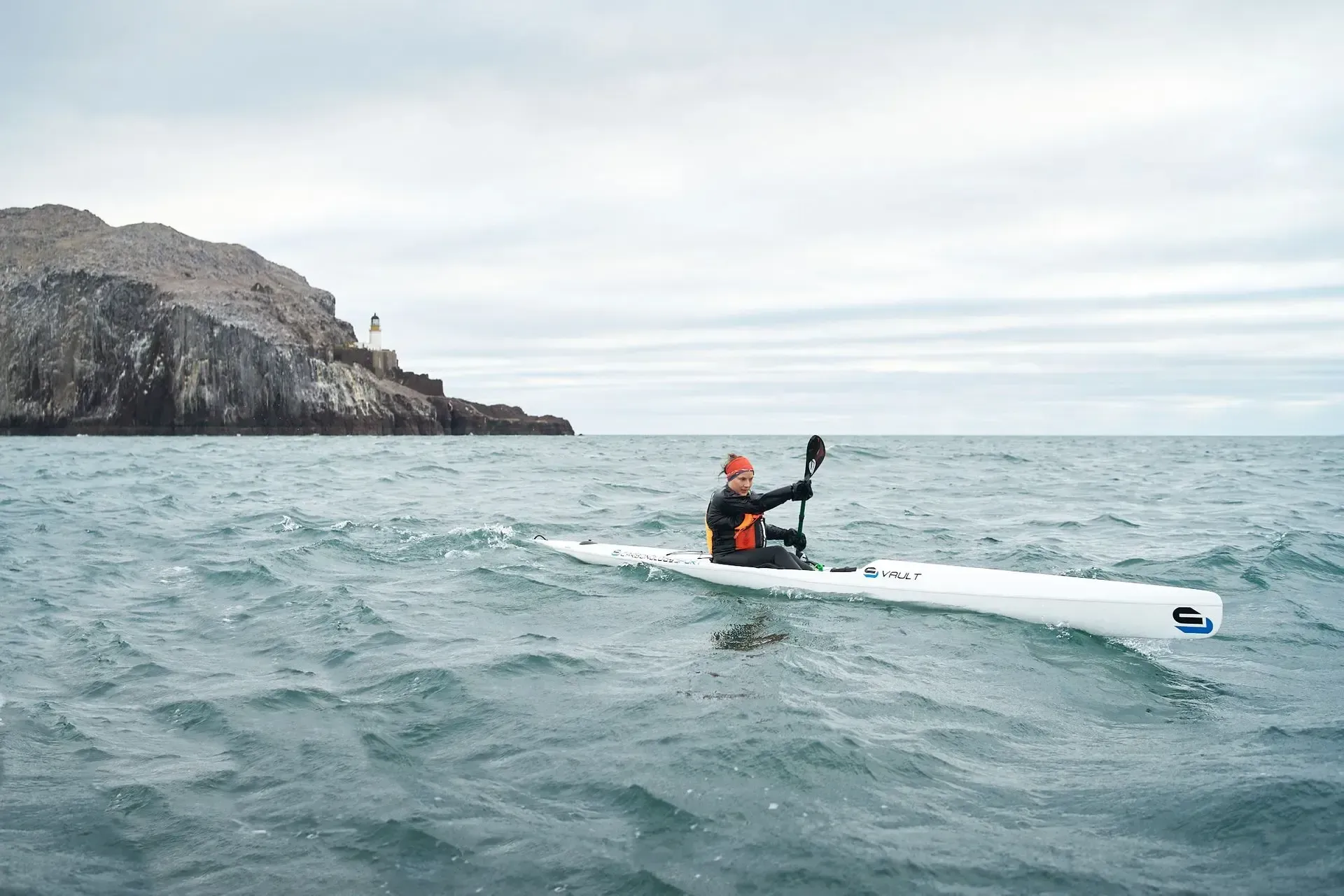Kayaker paddling in rough, grey water with a rocky island and overcast sky in the background.