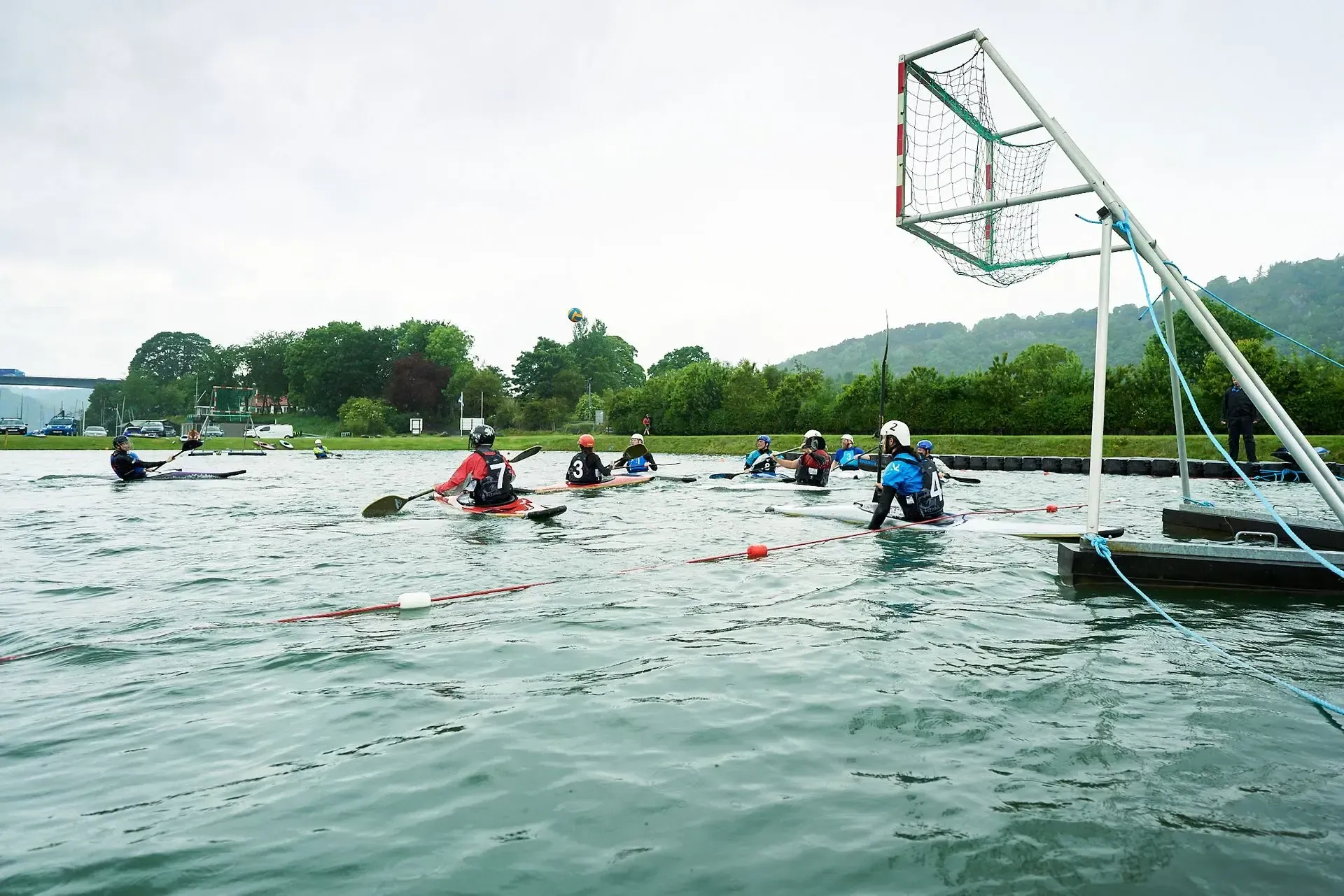 Kayakers playing water polo in a lake, aiming for a net structure. Cloudy day.