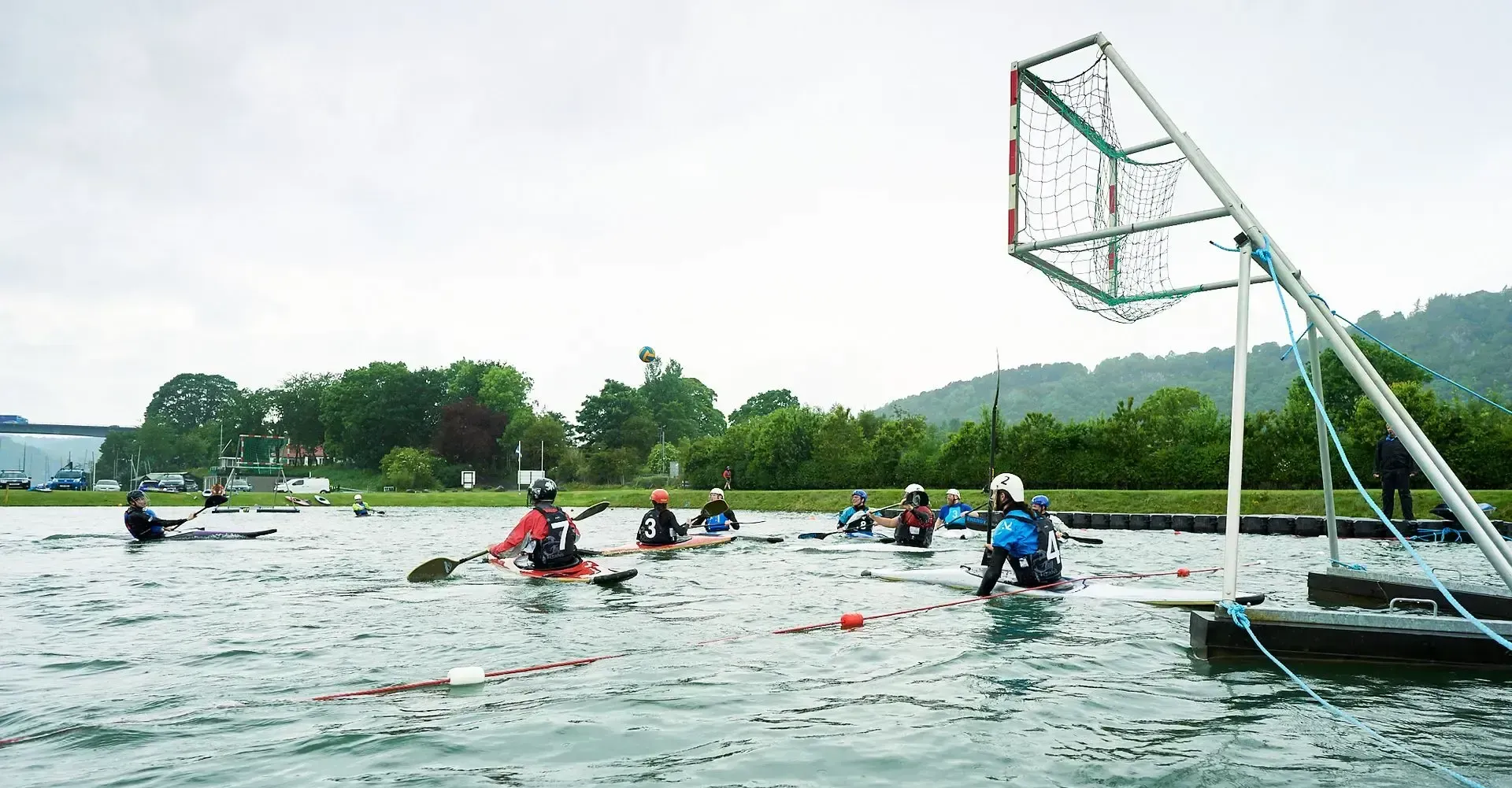 Kayakers playing water polo in a lake; a net is positioned above the water. Cloudy sky, trees in background.