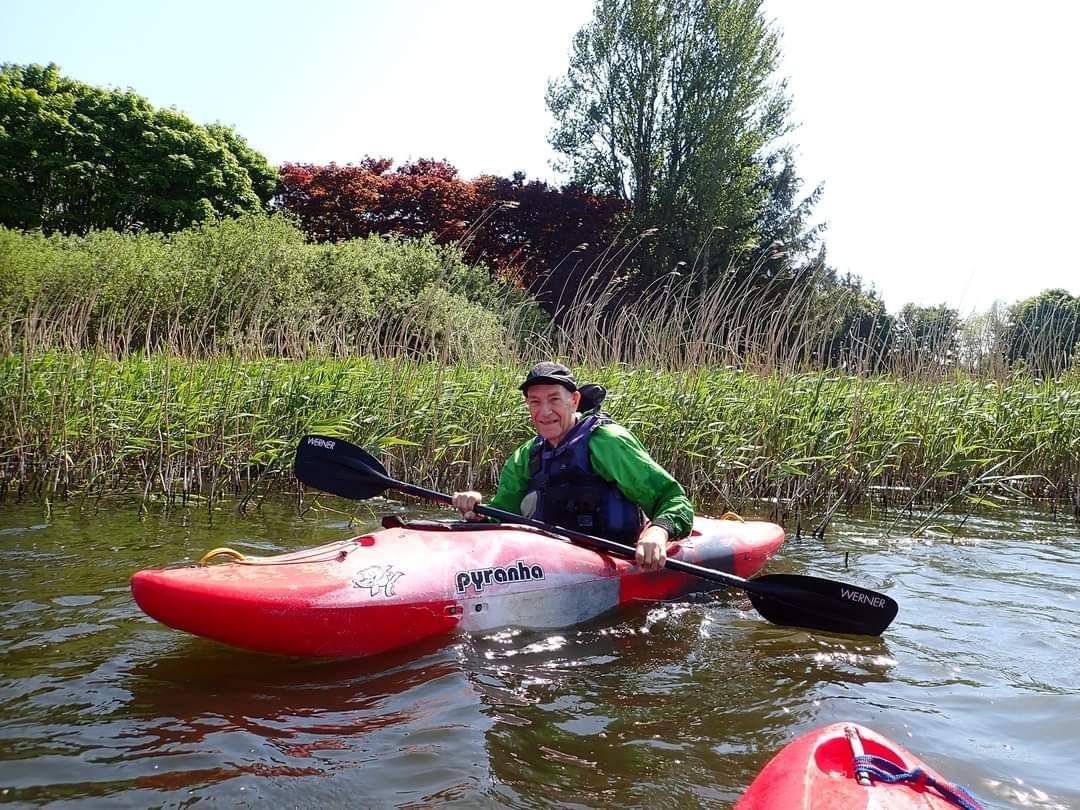 Person kayaking in a red and white kayak on calm water, surrounded by green foliage. The person wears a green jacket and smiling.
